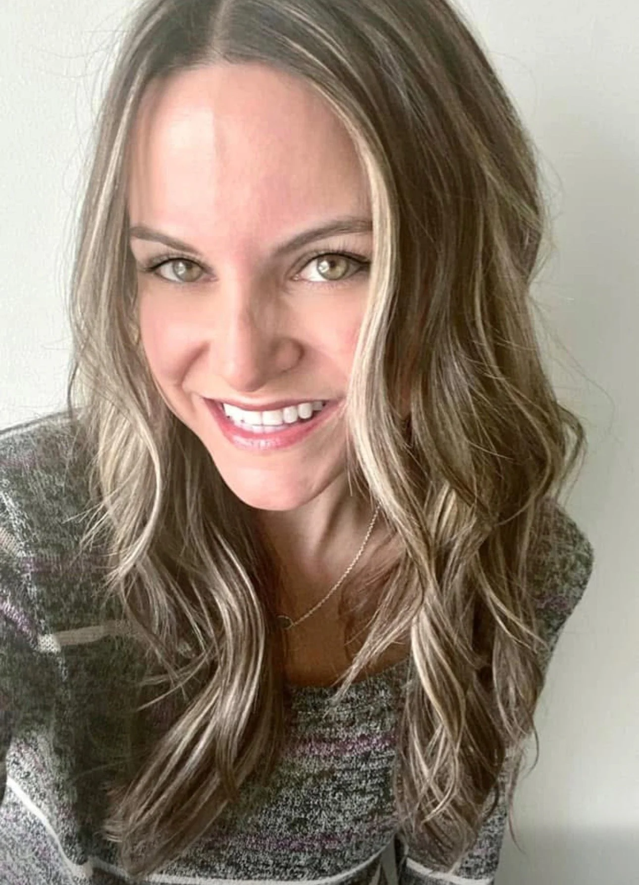 Close-up of a smiling woman with long wavy hair and light-colored eyes, wearing a patterned top and a delicate necklace.
