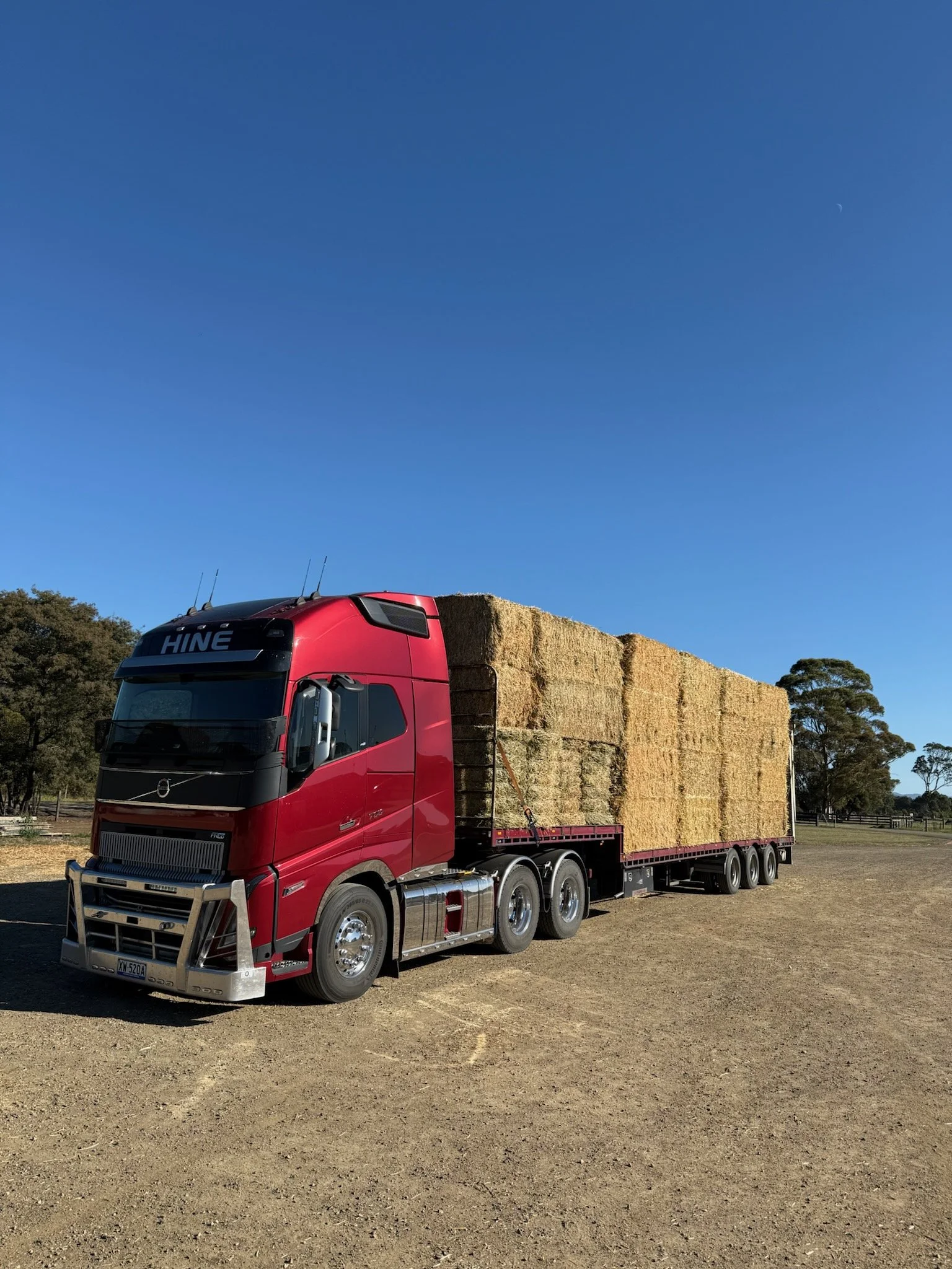 Red semi-truck loaded with a large stack of hay bales parked on a dirt field under a clear blue sky.