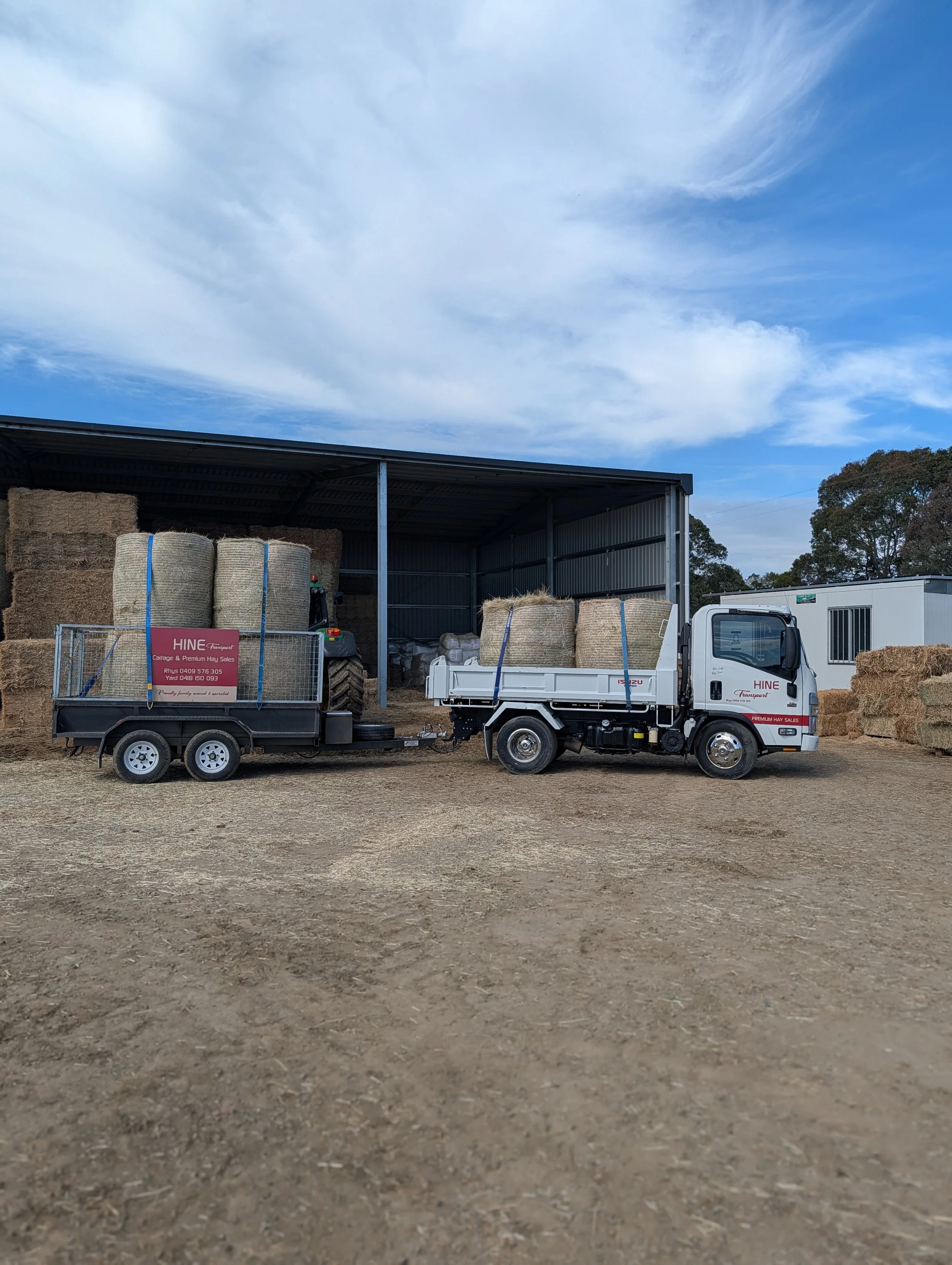 A white truck with a flatbed loaded with hay bales, parked in front of a large open-sided shed filled with more hay bales, on a dirt ground under a cloudy sky.