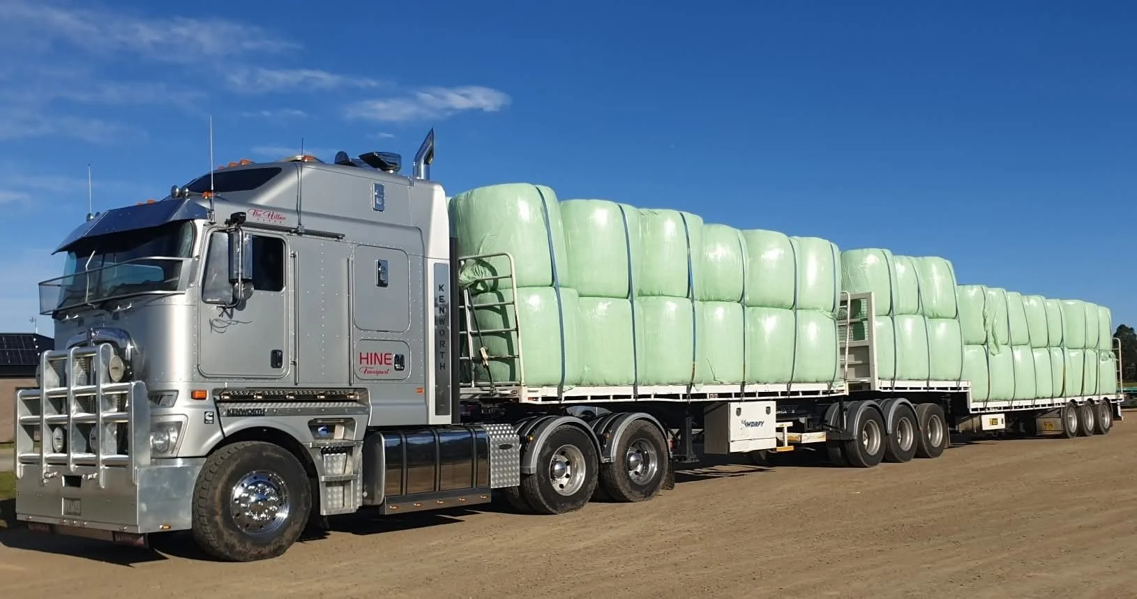 A large semi-truck carrying multiple large green bales of hay on a dirt road under a bright blue sky.