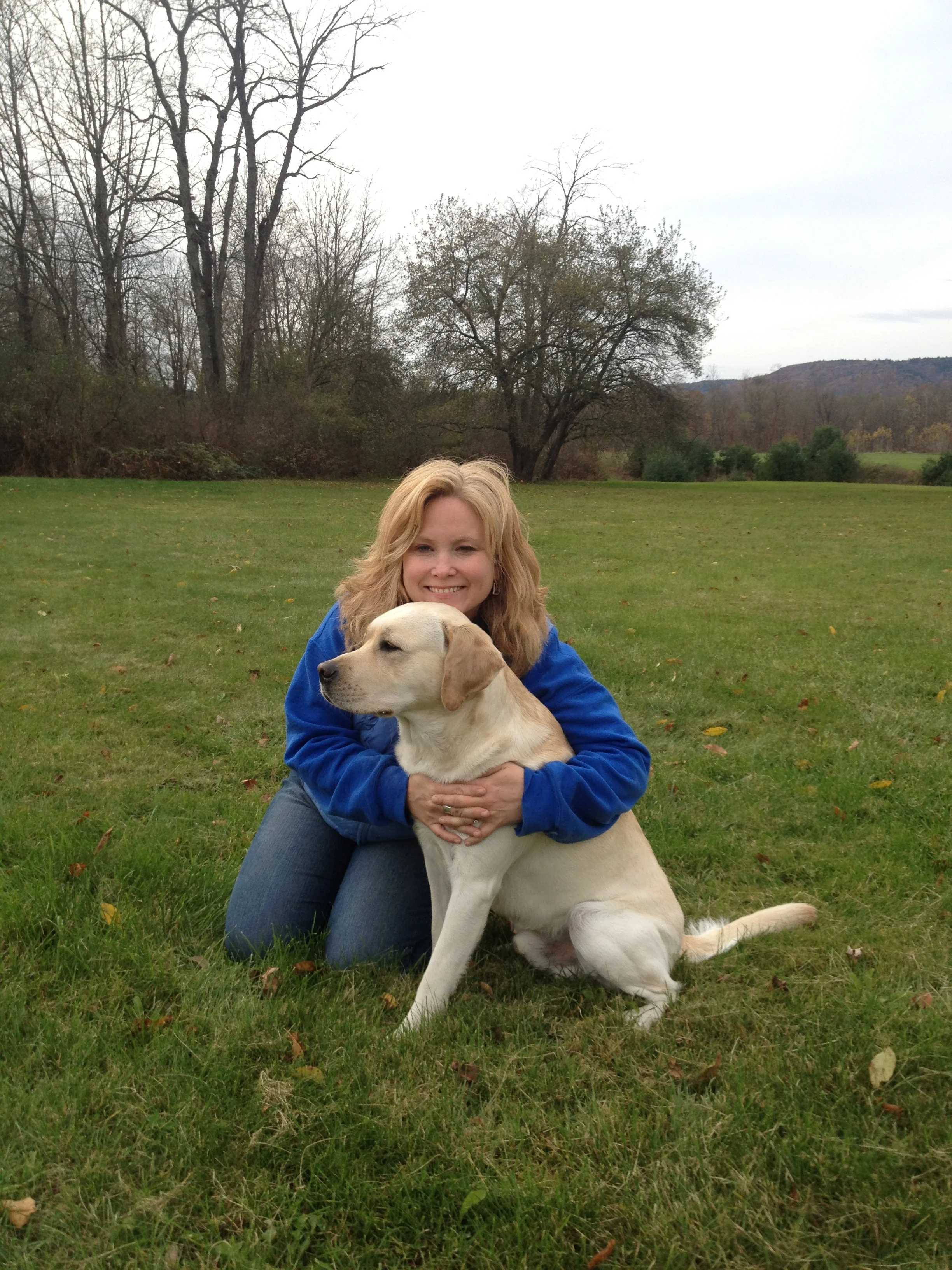 A woman with blonde hair wearing a blue jacket sitting on grass and hugging a yellow Labrador retriever dog outdoors in a park or field with trees and hills in the background.