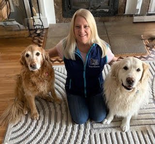 A woman with blonde hair wearing a blue vest, sitting on a striped rug, smiling, with a golden retriever and a Labrador retriever sitting next to her indoors.