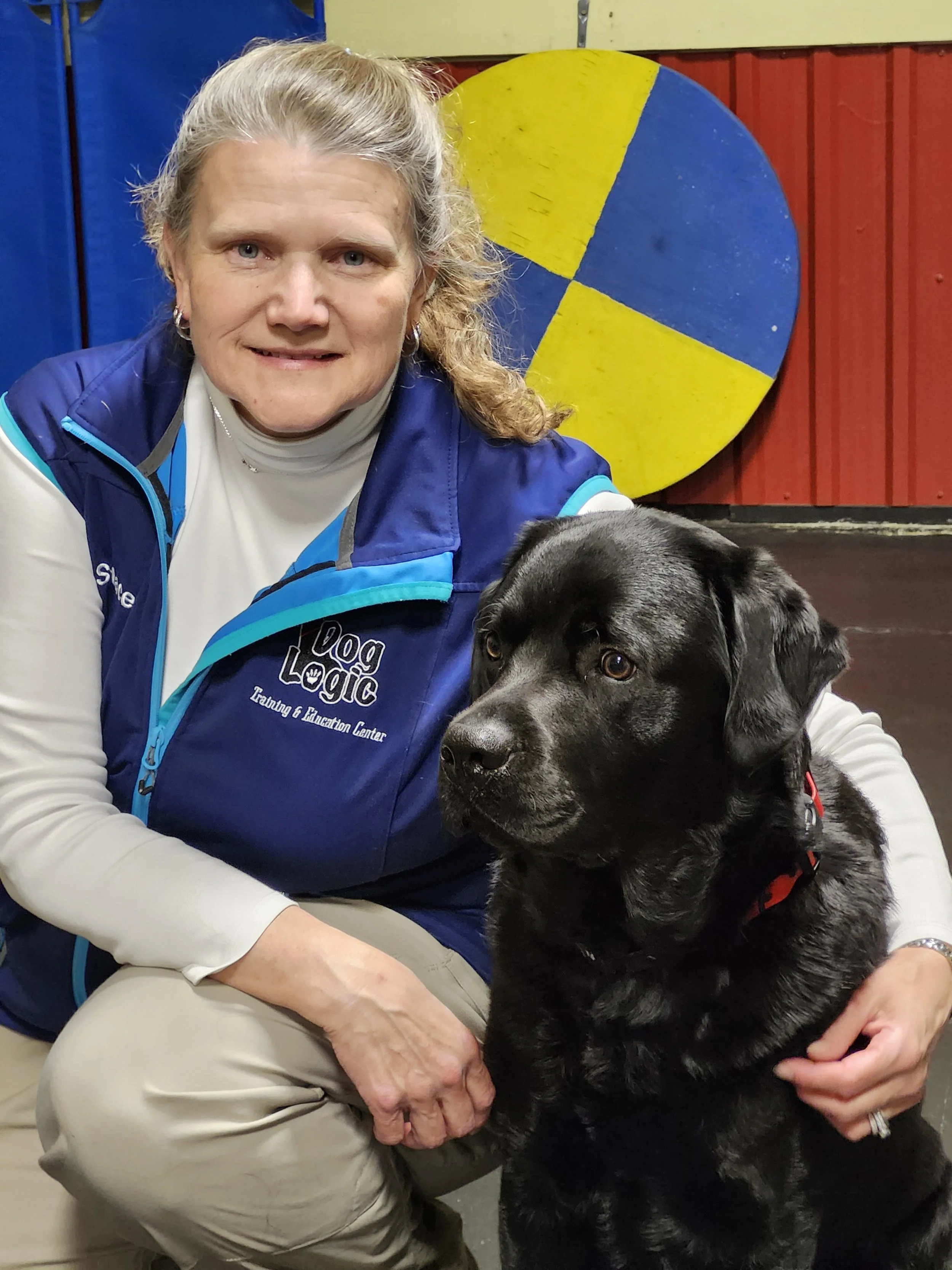 A woman wearing a blue vest with 'Dog Logic' text, and a white turtleneck, sitting indoors with a black dog that has a red collar. The woman is smiling and the dog appears calm, sitting next to her.