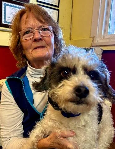 An elderly woman with glasses holding a small, fluffy, black and white dog indoors in front of yellow walls and framed certificates.