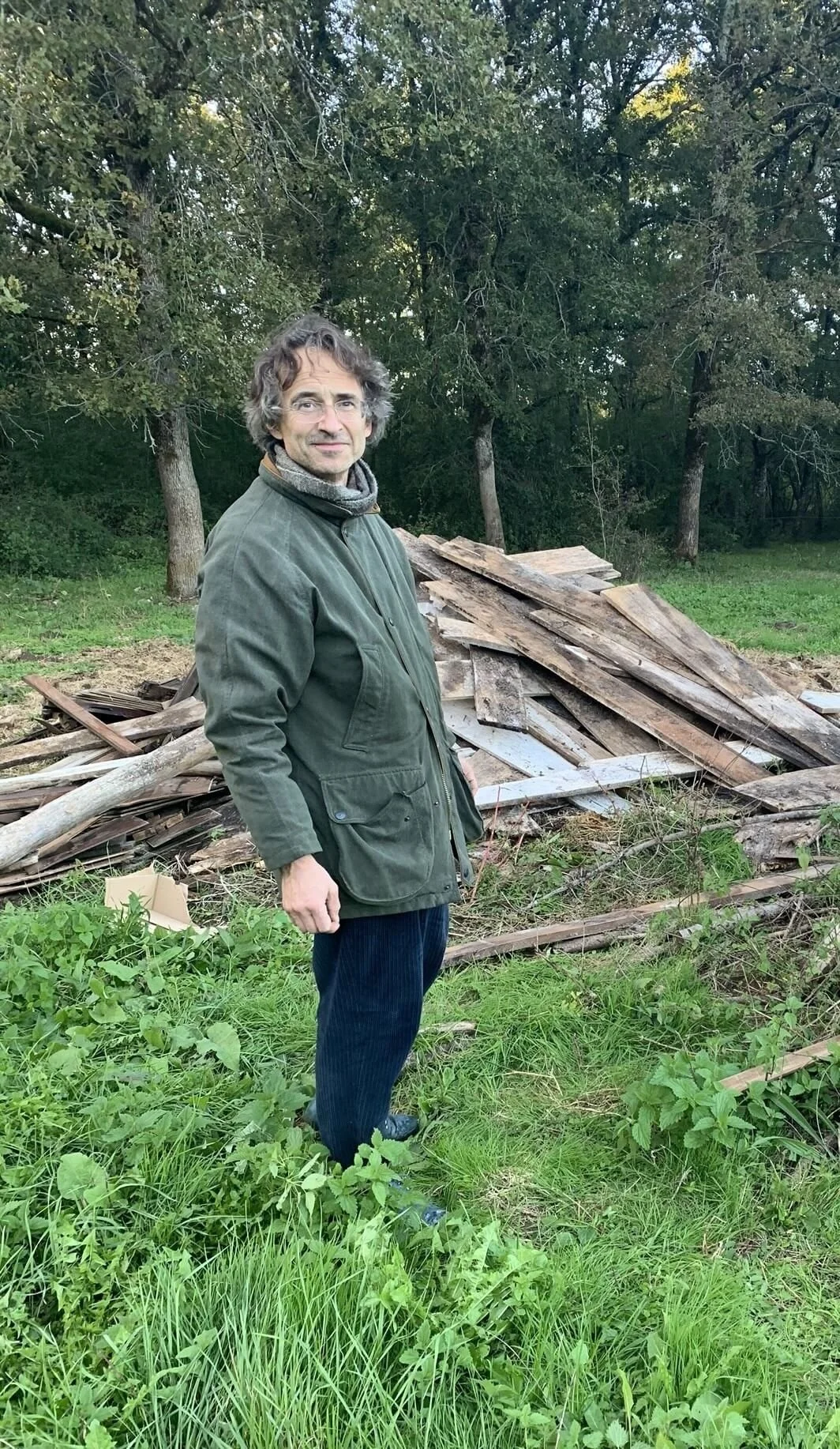 A middle-aged man with gray hair and glasses standing outdoors in a green landscape, wearing a scarf, jacket, and a slight smile.