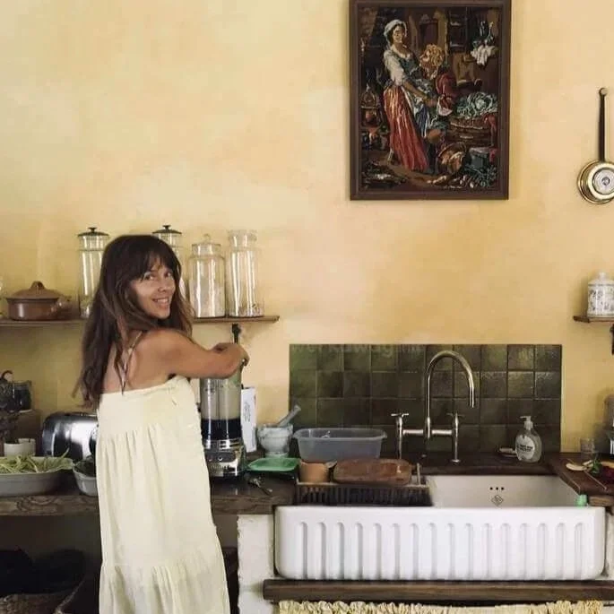 A woman with wavy, shoulder-length brown hair and blue eyes taking a selfie indoors, wearing a patterned top and a turquoise necklace.