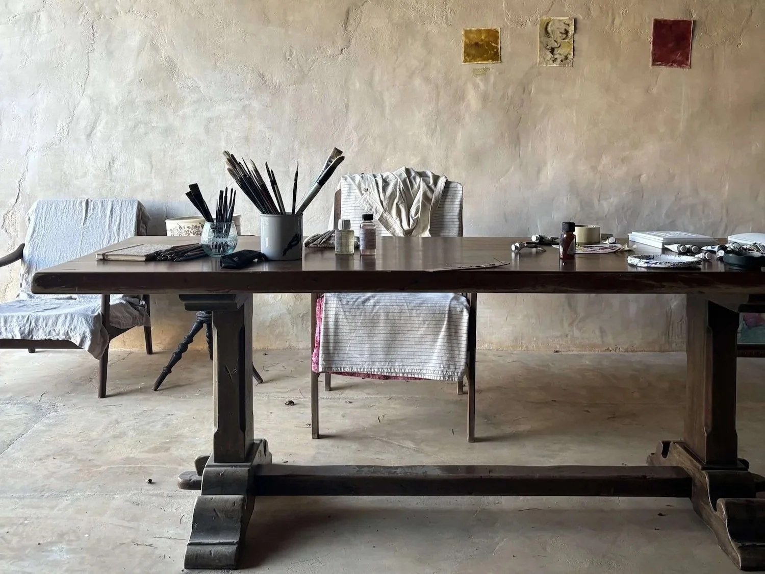 A large wooden table with various art supplies, including brushes in containers, jars, and bottles. Two chairs with cloth covers and a wall with three small decorative tiles in the background.