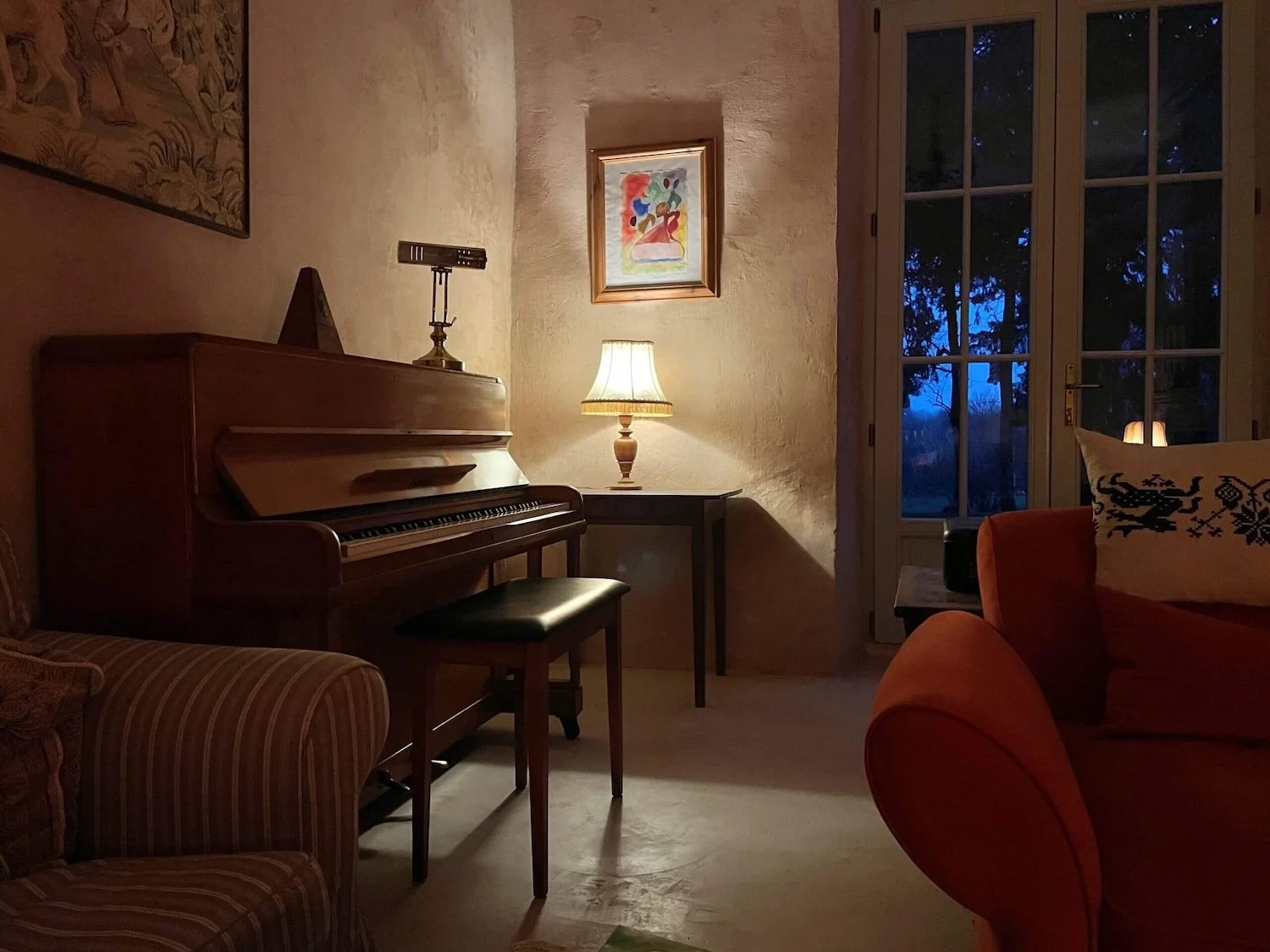 A living room with a wooden upright piano, a side table with a lit table lamp, a framed colourful artwork on the wall, a large window with blue evening sky and trees outside, a burnt orange armchair with a decorative pillow, and part of a striped 