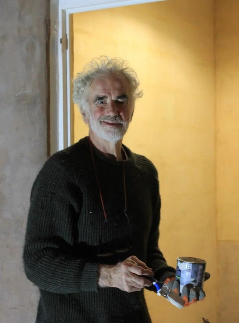 Close-up of an older man with white hair, beard, and glasses, wearing a brown leather jacket with a shearling collar.