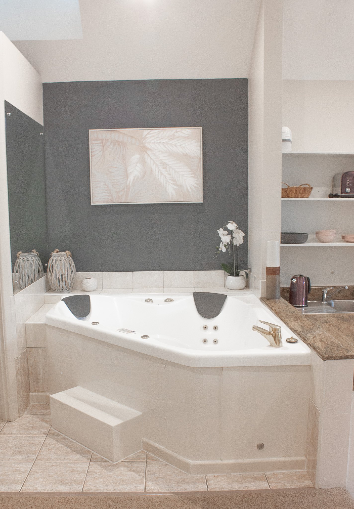 A bathroom corner featuring a white bathtub with black headrests, a dark wall behind, a framed artwork, and decorative vases. Open shelves with various items are to the right.