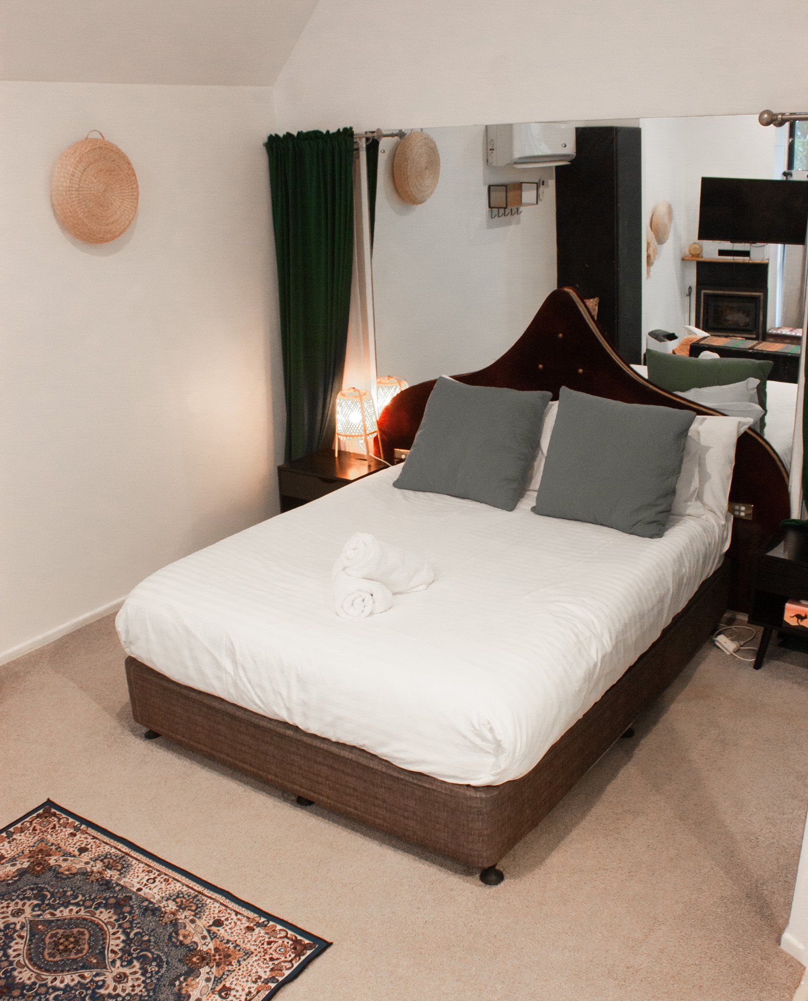 A bedroom with a bed, gray pillows, a towel folded into a swan, a nightstand with a lamp, a mirror, green curtains, and decorative wall hangings.