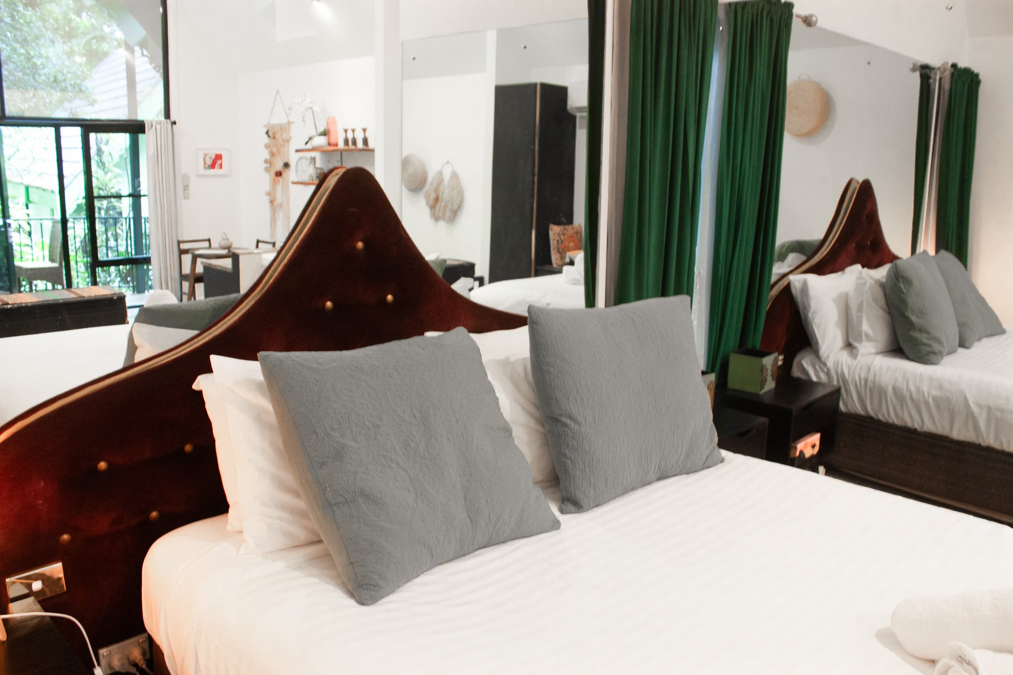 Two beds with dark wooden headboards, each with gray pillows and white linens, in a well-lit hotel room with large windows, green curtains, and decorative wall hangings.