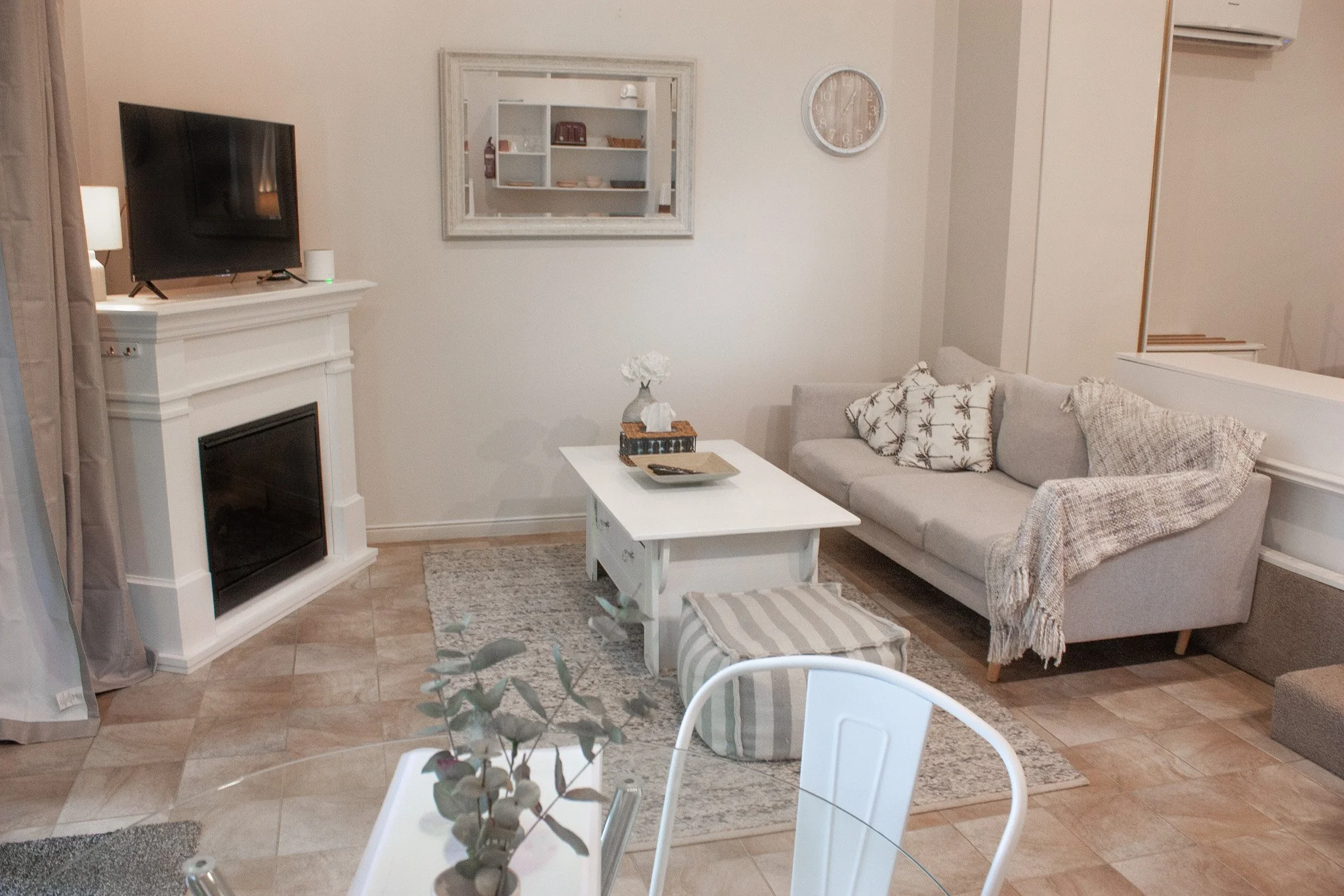 Living room with white walls, beige tiled floor, a white coffee table, a gray sofa with patterned pillows, a fireplace with a TV on top, a mirror on the wall, and a round wall clock.