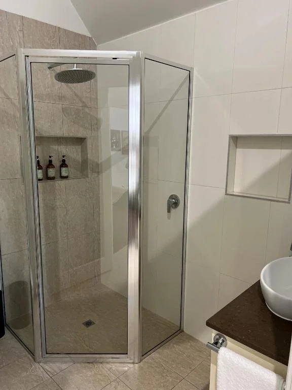 Bathroom shower area with glass enclosure, built-in shelf with bottles, and a white vessel sink on a dark countertop.