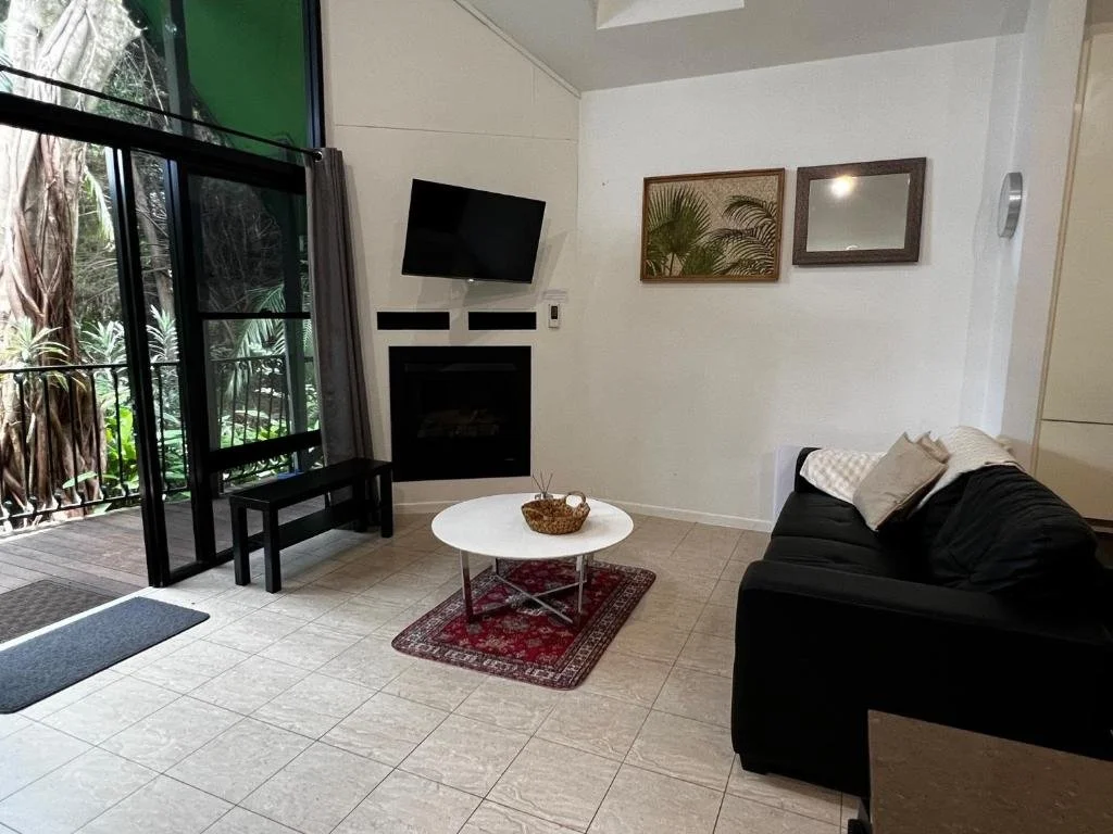 Living room with black sofa, white pillows, wall-mounted TV, fireplace, wall art, glass door leading outside, small bench, round white coffee table, red patterned rug, green framed mirror, and forest visible outside.