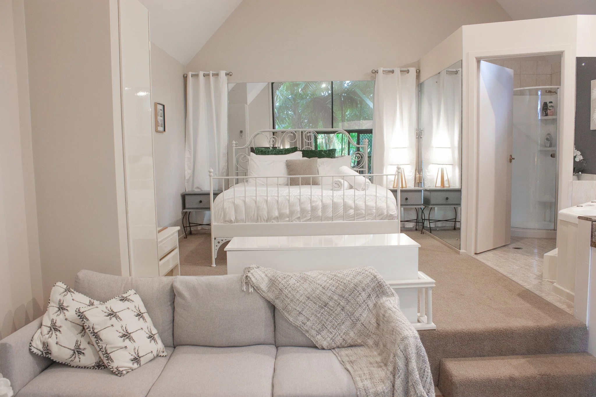 A cozy bedroom with a white bed, white curtains, and a large window with greenery outside. There are two nightstands with lamps, a white wardrobe, and a mirror. The room opens into a bathroom with tiled floors, and a light-colored sofa with pillows a