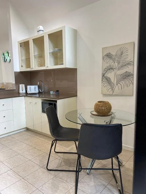 A modern kitchen with white cabinets, a brown countertop, and a circular glass dining table with two black chairs. There's a wall art piece featuring palm leaves and decorative items on the table, along with kitchen appliances on the counter.