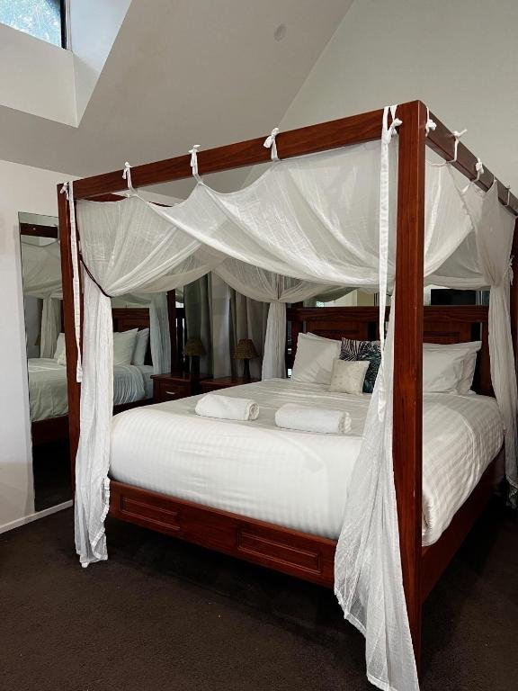 A four-poster bed with a white canopy and drapes, made with white bedding and pillows, a folded towel, in a hotel room with a mirror reflecting part of the bed.