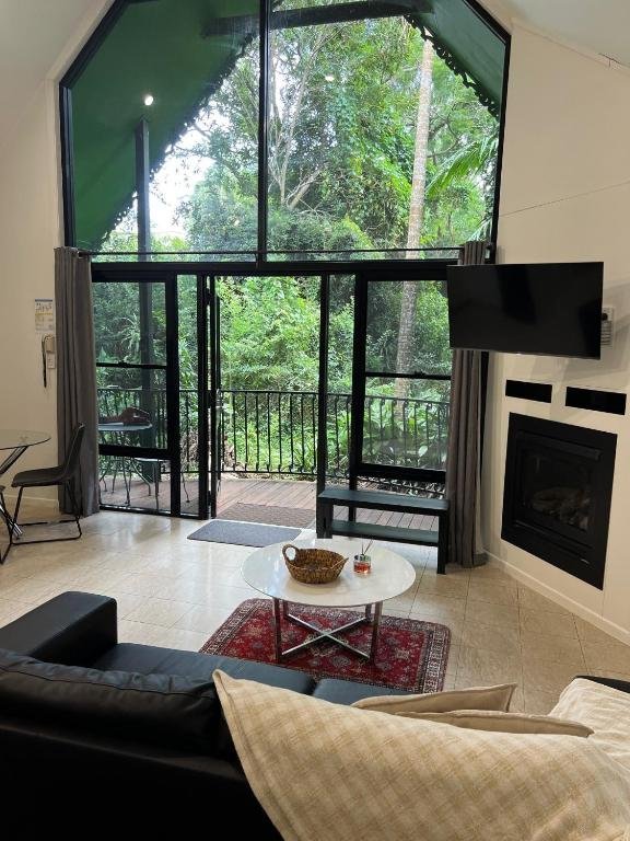 Modern living room with large window and glass door leading to a balcony overlooking lush greenery, a wall-mounted TV, a fireplace, a dark leather sofa with beige cushions, a round white coffee table with a basket and a drink, a small red rug underne