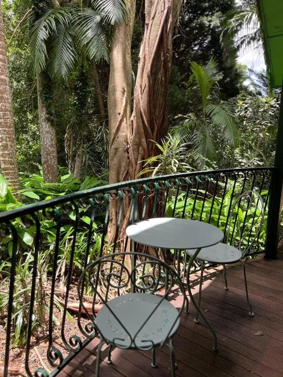A small outdoor balcony with a black decorative metal railing, three white metal chairs, and a small round table. The balcony overlooks lush green tropical plants and trees.