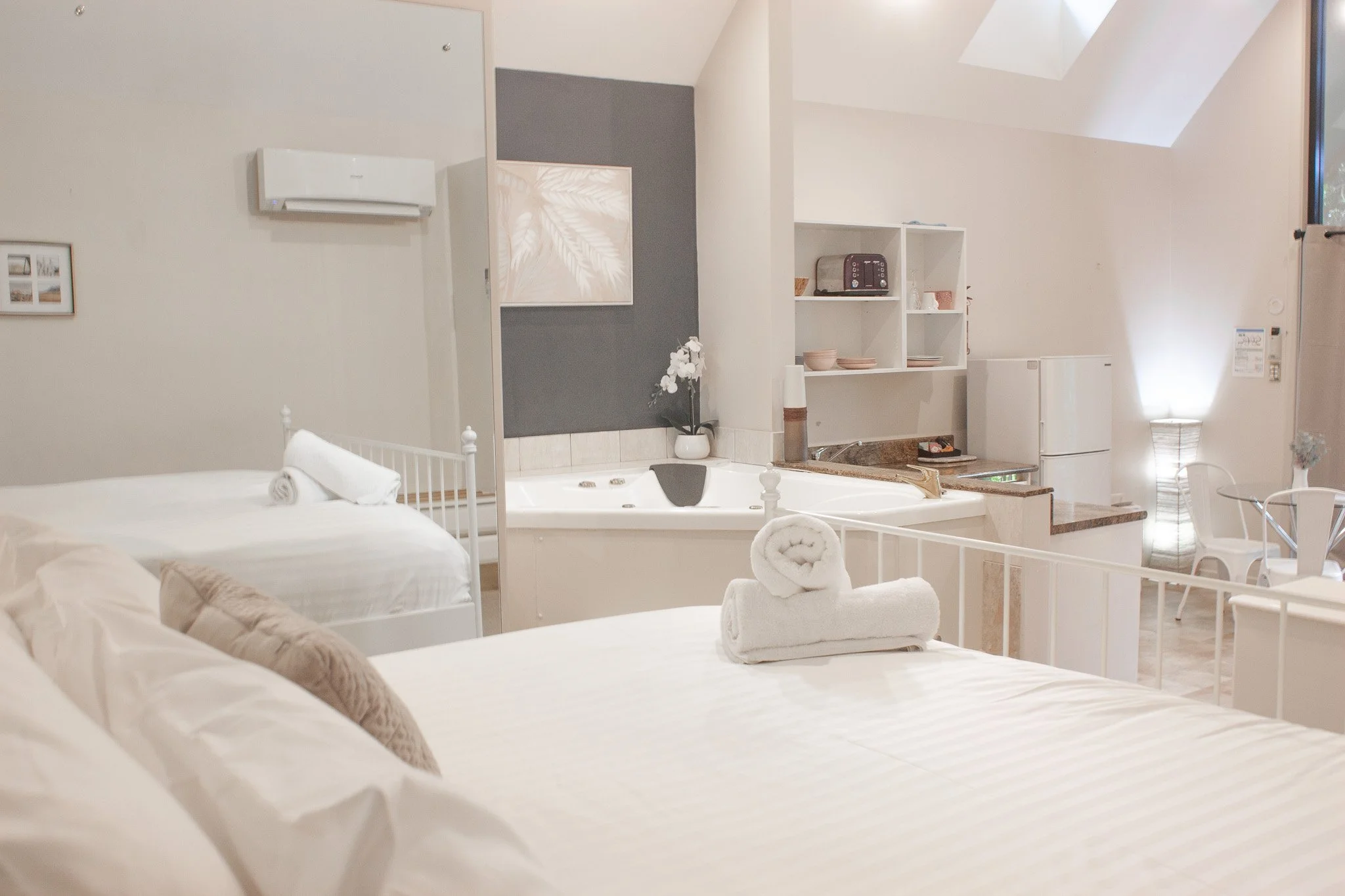 Hotel room with two white beds, one with rolled towels, a Jacuzzi, kitchenette, dining table, and a dark accent wall with framed artwork.