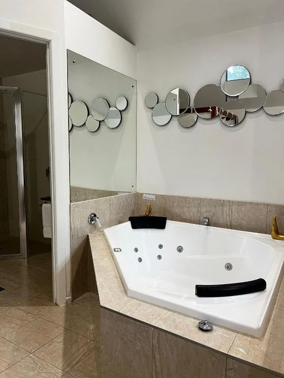 A corner bathtub with black headrests and gold fixtures in a bathroom with beige tiles and circular mirrors on the wall.