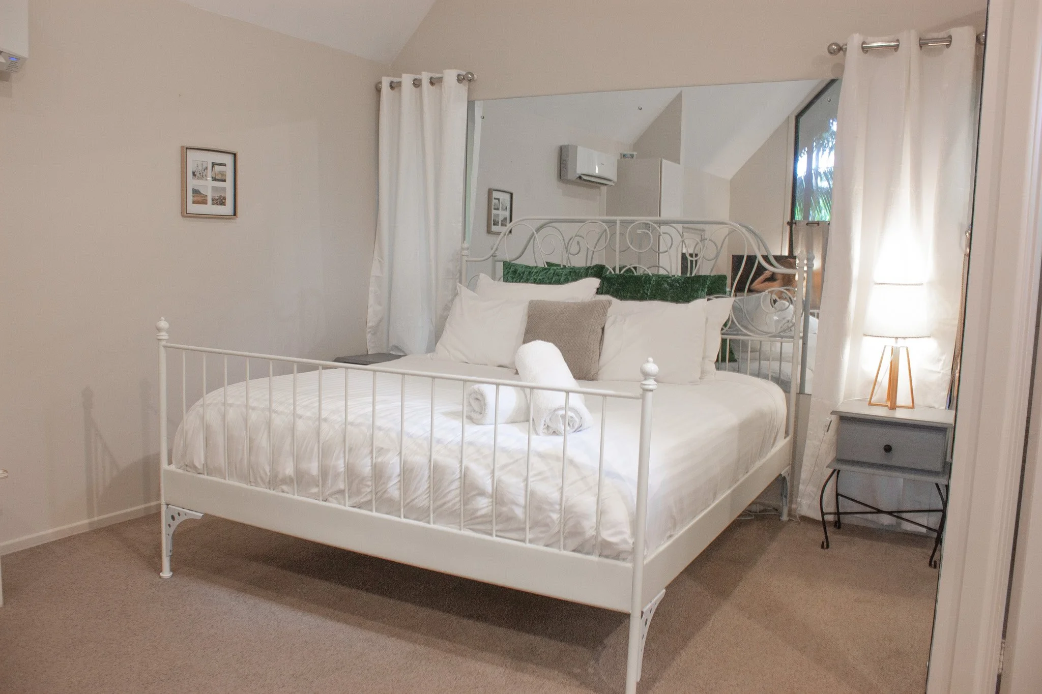 Neatly made bedroom with a white metal bed frame, white bedding, rolled towels, pillows, green decorative pillows, a side table with a lamp, and white curtains.
