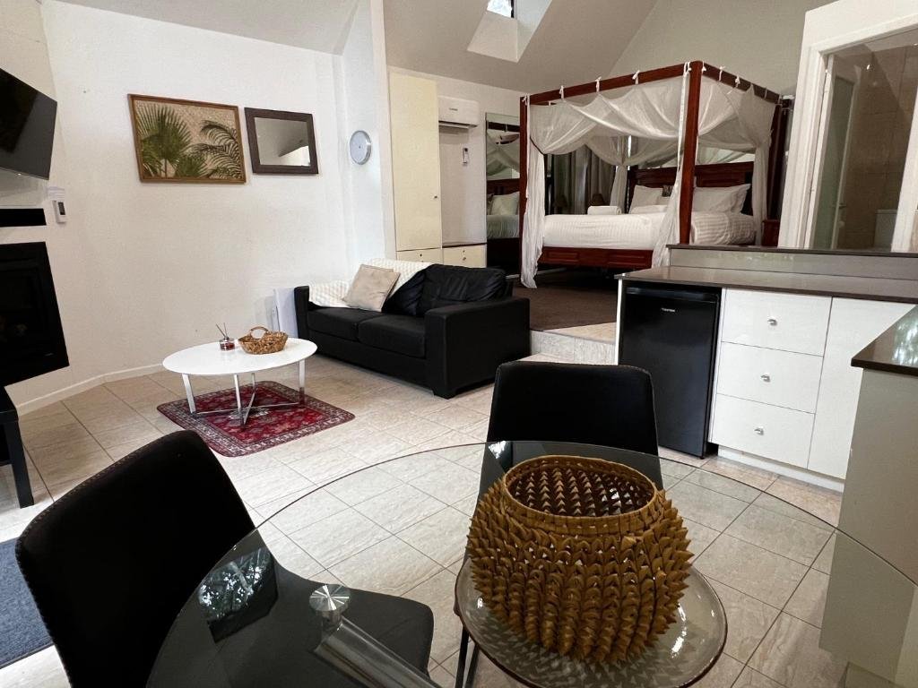 A cozy studio apartment with a dining area, black sofa, white coffee table on a red rug, a queen-sized bed inside a canopy frame, and white walls with two framed pictures and a mirror.