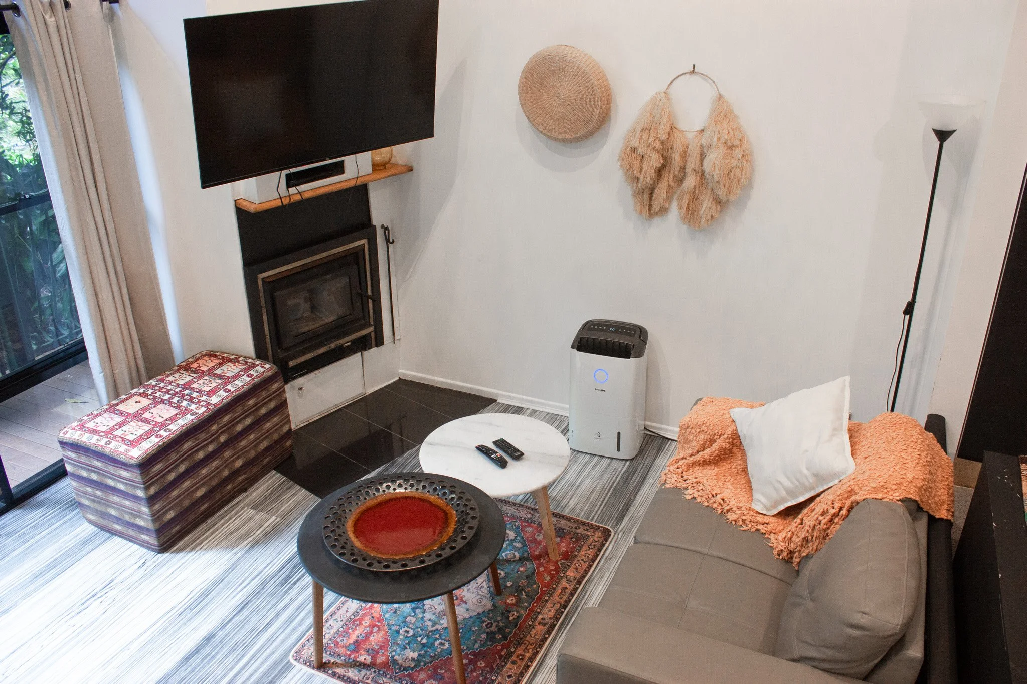 A cozy living room with a large flat-screen TV on the wall above a fireplace, a cushioned bench with patterned upholstery, a small round coffee table with remote controls, a sofa with an orange throw blanket and a white pillow, a dehumidifier, a tall