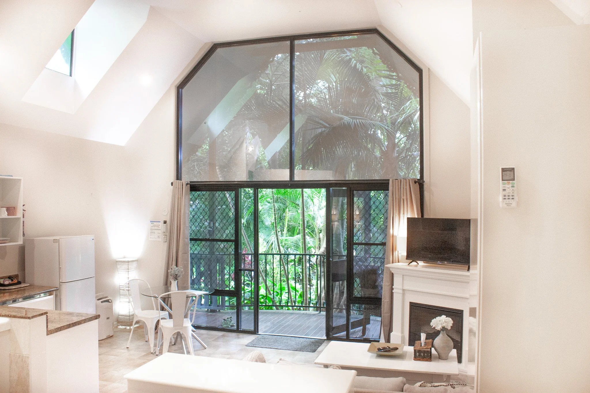 Living room with large glass windows and doors leading to a balcony, overlooking a lush green tropical garden. The room has a white fireplace, a small TV on top, and a white table with decorative items and flowers.
