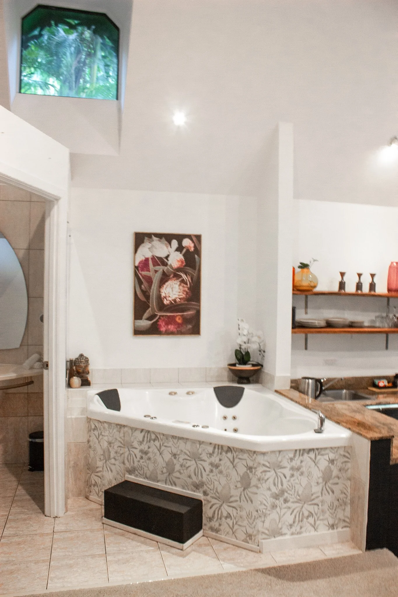 A bathroom with a white corner whirlpool bathtub decorated with a floral tile pattern. There is a black step stool in front of the tub, and a small green potted plant on a ledge nearby. The room features white walls, a wooden shelf with dishes and de