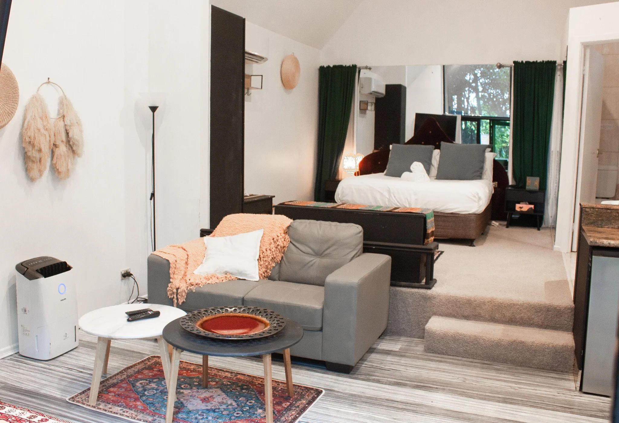 Studio apartment with a living area and bedroom, featuring a gray couch with a peach blanket, two small round coffee tables, a bed with white and gray pillows, a window with green curtains, and various decorative items.