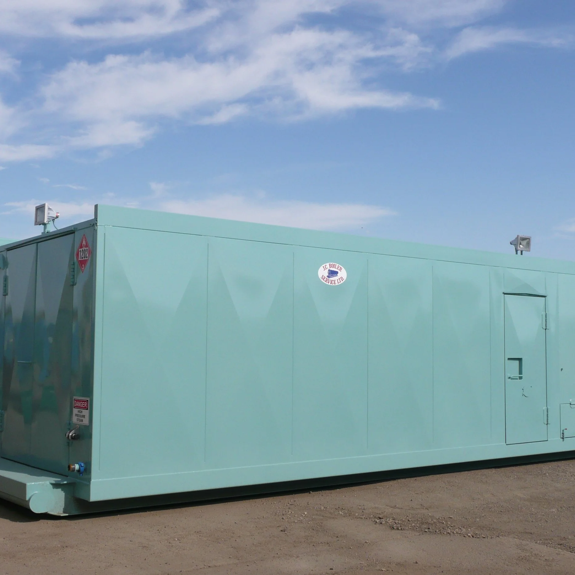 A large, mint green industrial boiler with warning labels and safety equipment, situated outdoors on a dirt surface, with a blue sky and wispy clouds overhead.