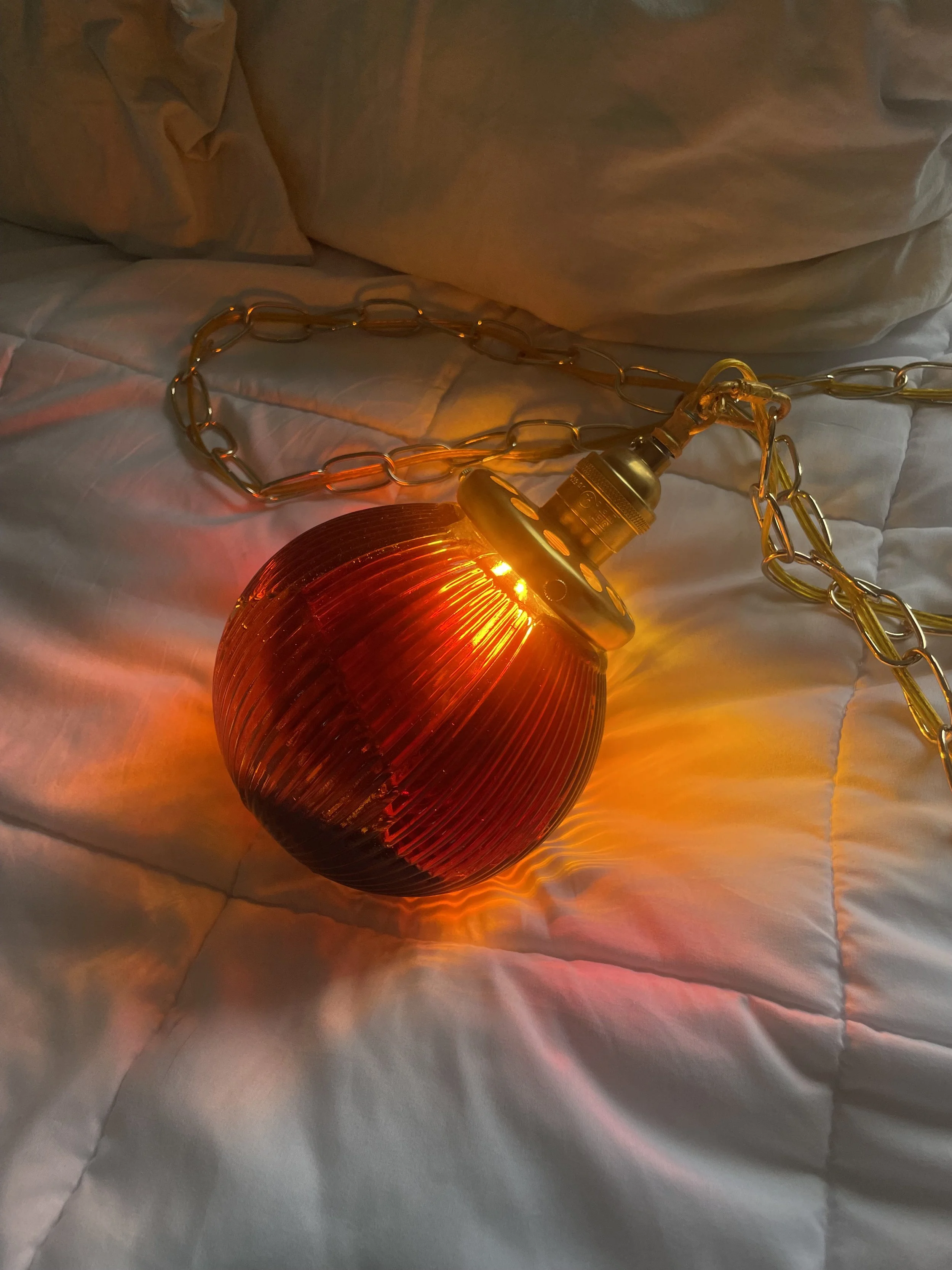 A vintage red resin pendant light with a gold chain lying on a white bedspread, emitting a warm glow.