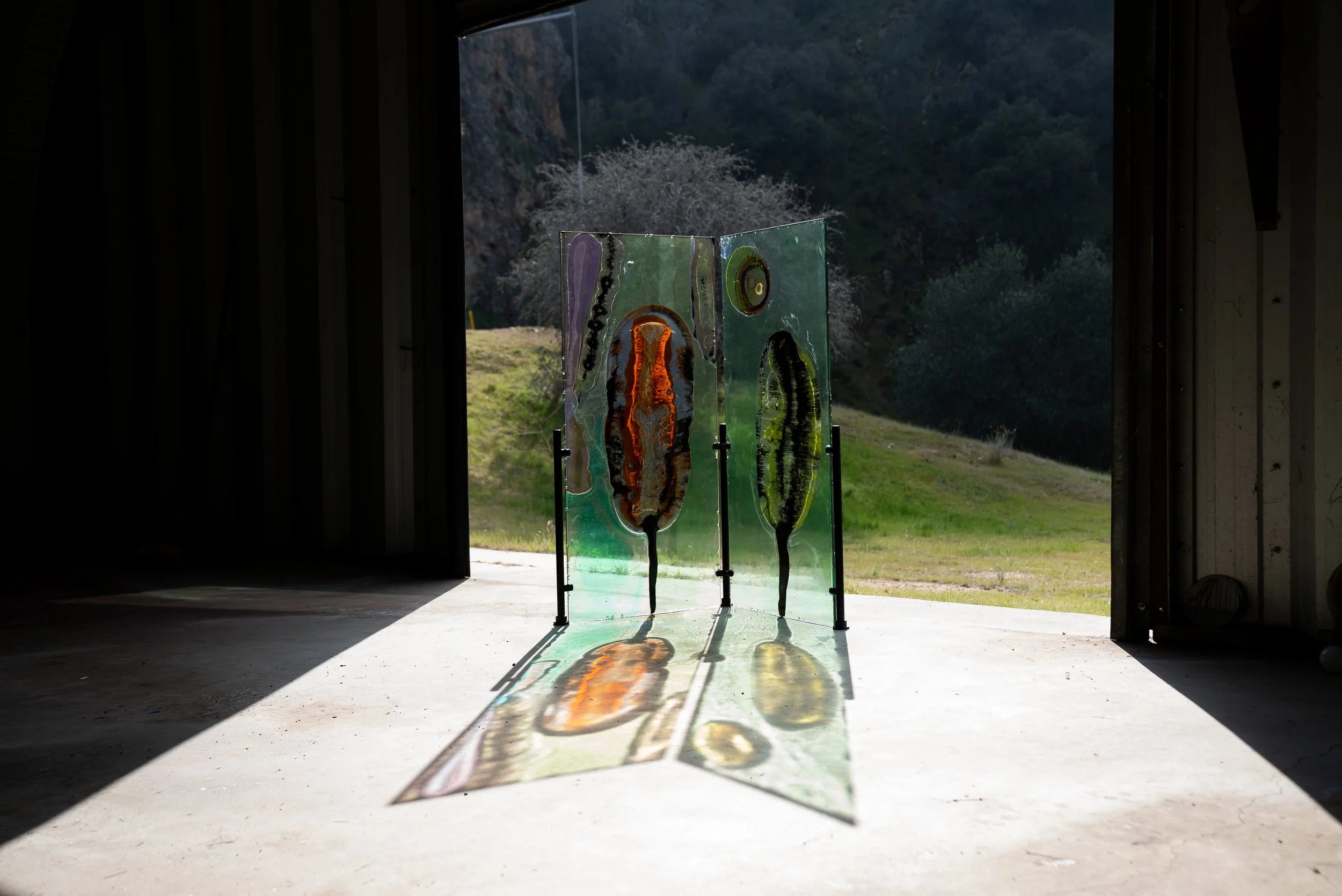 A resin art installation with colorful, abstract patterns stands inside a dark barn, with an open doorway revealing a grassy landscape and hills outside.