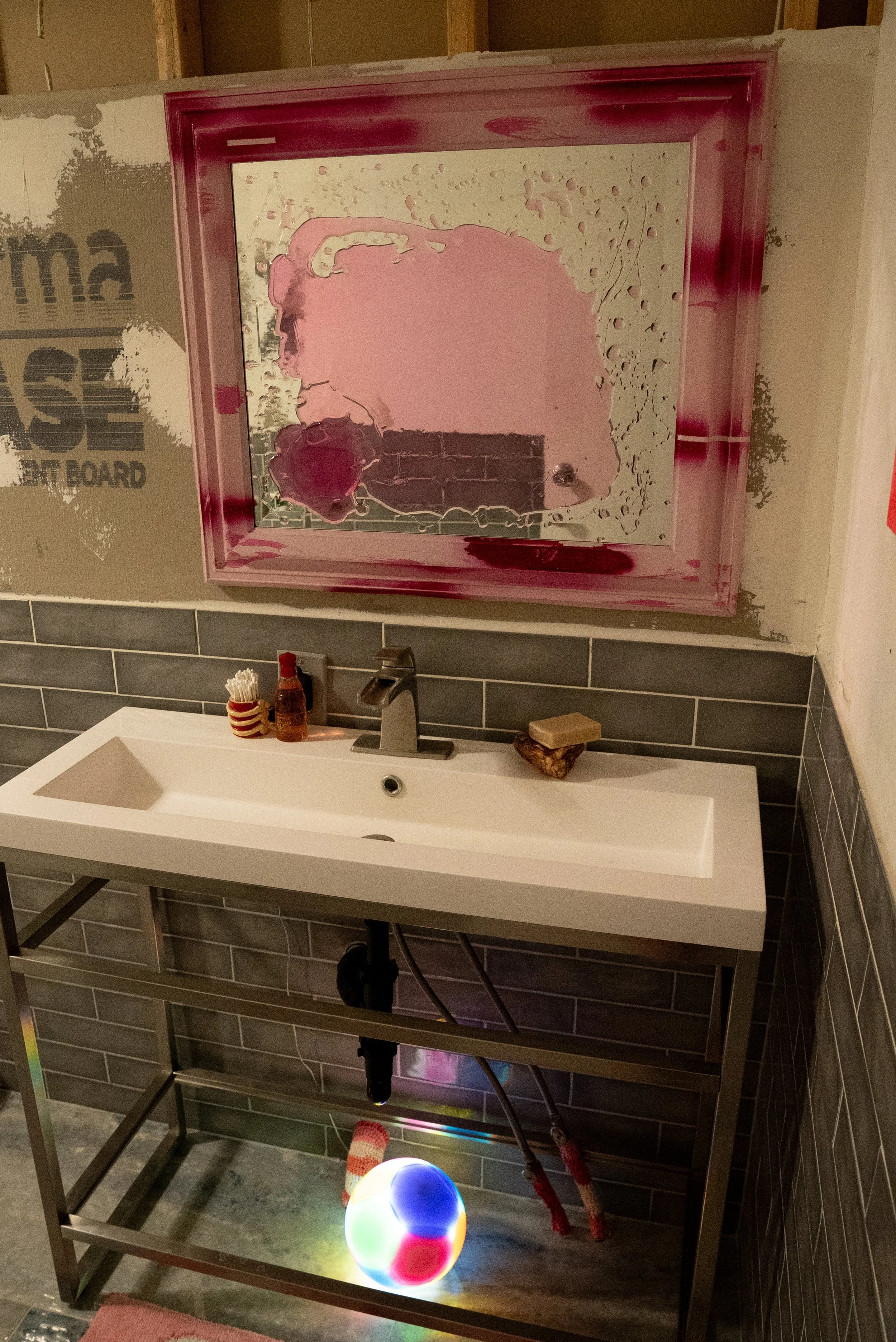 A pink resin mirror with pink frame above a rectangular white sink. A multicolored glowing resin ball is on the floor beneath the sink, and the wall has grey tiles and unfinished drywall.