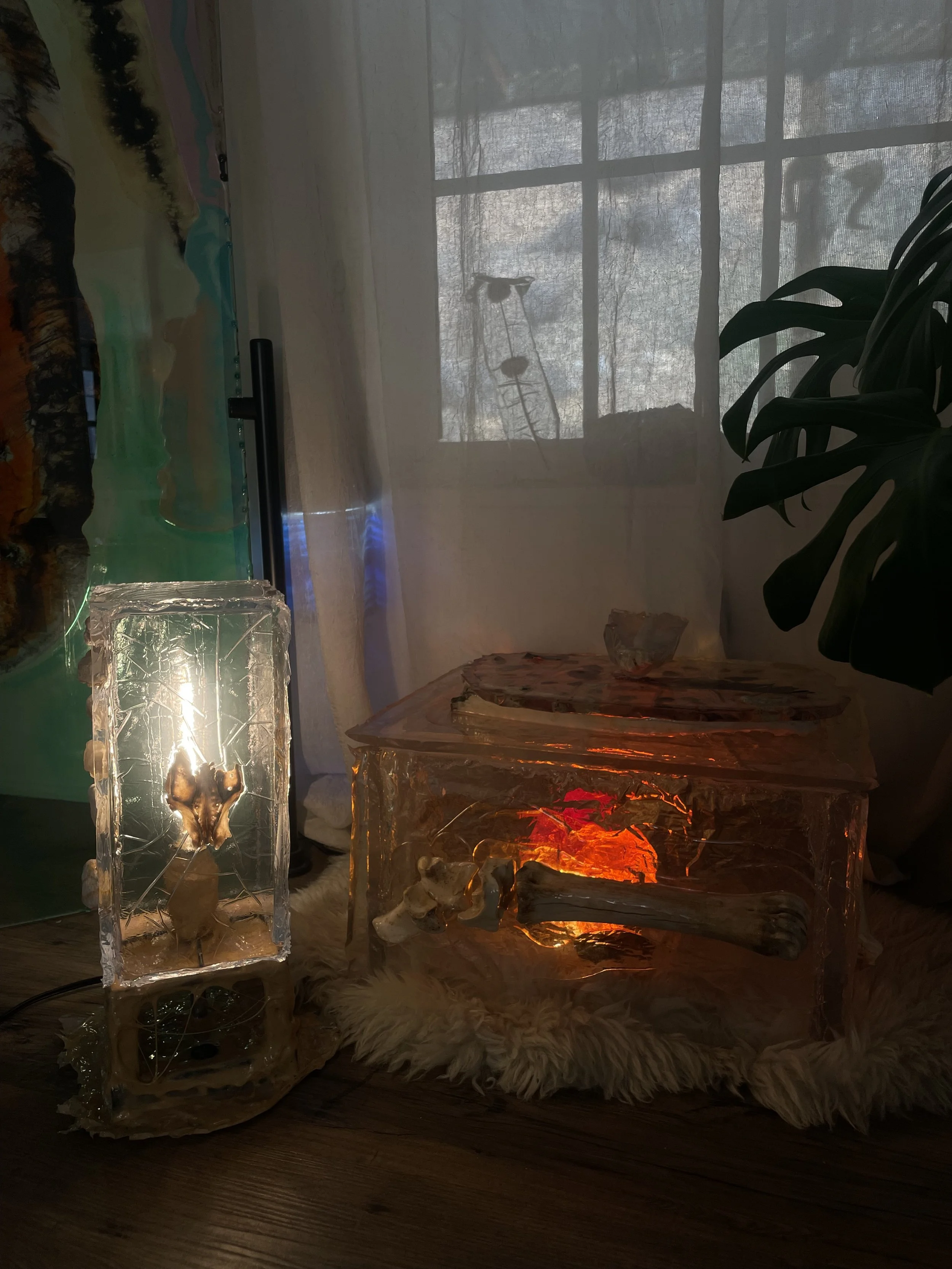 Decorative ice or resin table and lamp with natural elements inside, placed on a fluffy rug near a window with sheer curtains and a leafy plant nearby.