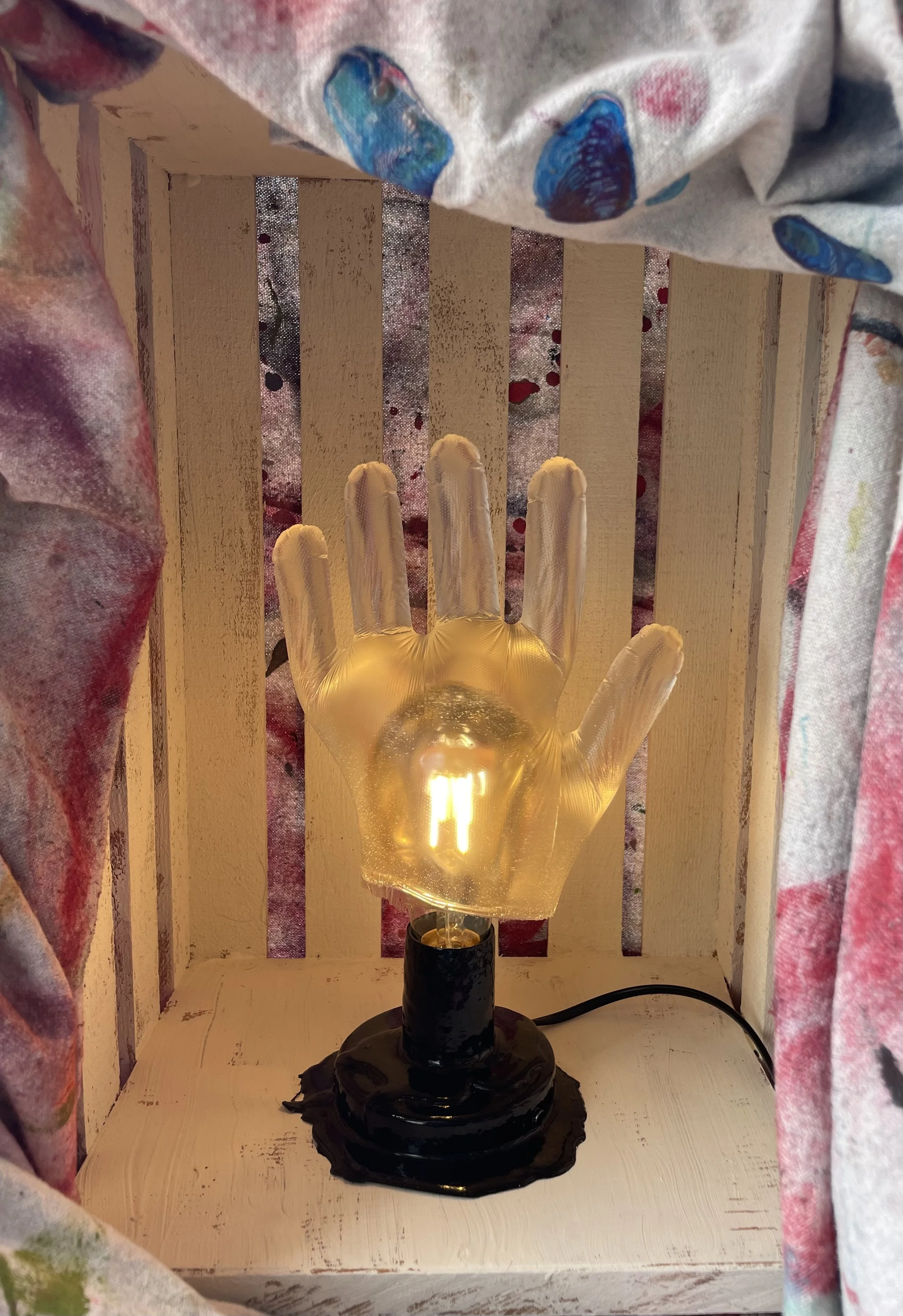 A hand-shaped resin lamp with a glowing light bulb inside, placed on a wooden surface with a white painted finish, surrounded by colorful curtains.