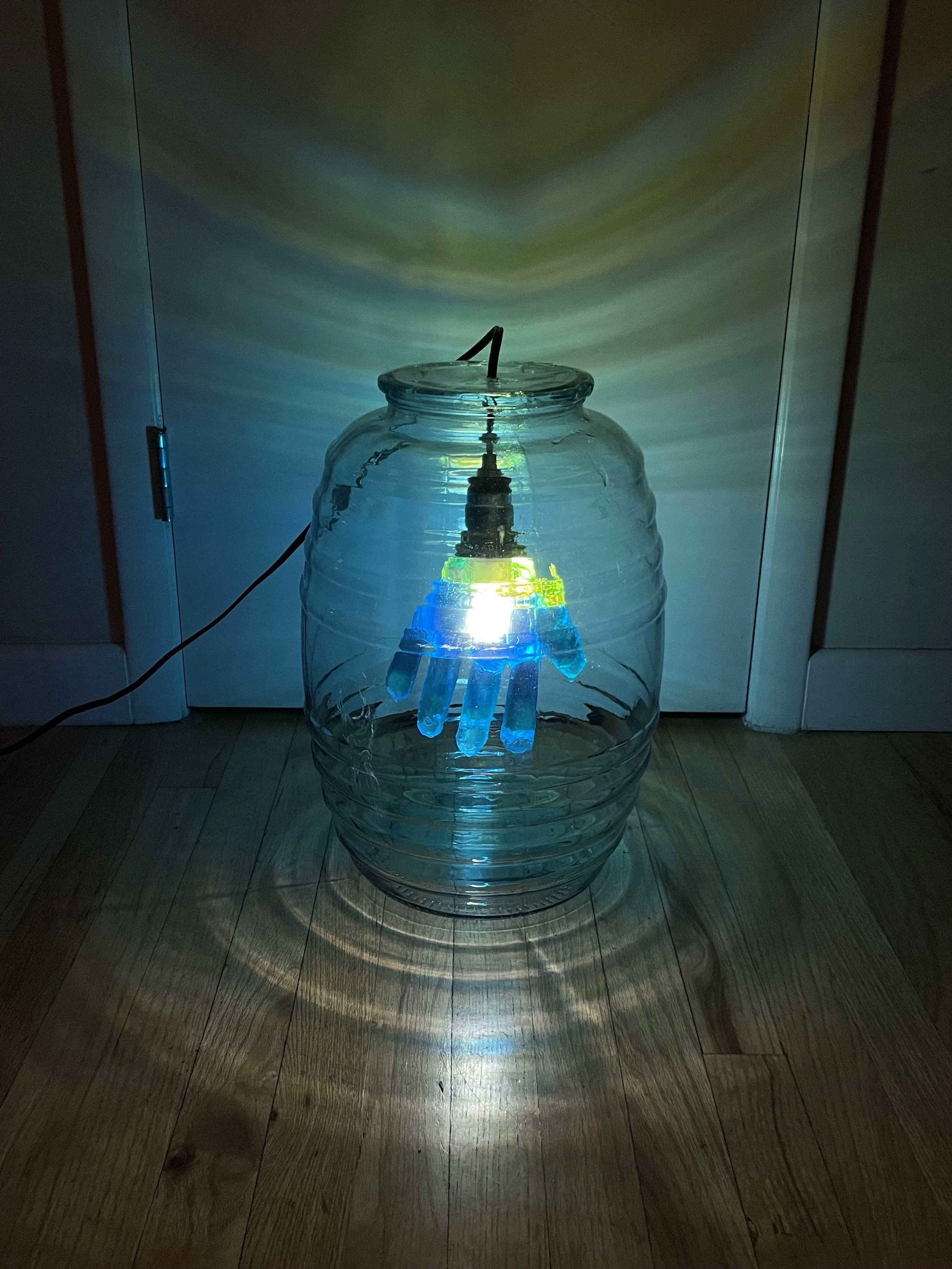 A large transparent glass jar with a light inside, illuminating a blue and yellow resin glove hanging inside, creating colorful reflections and shadows on the floor.
