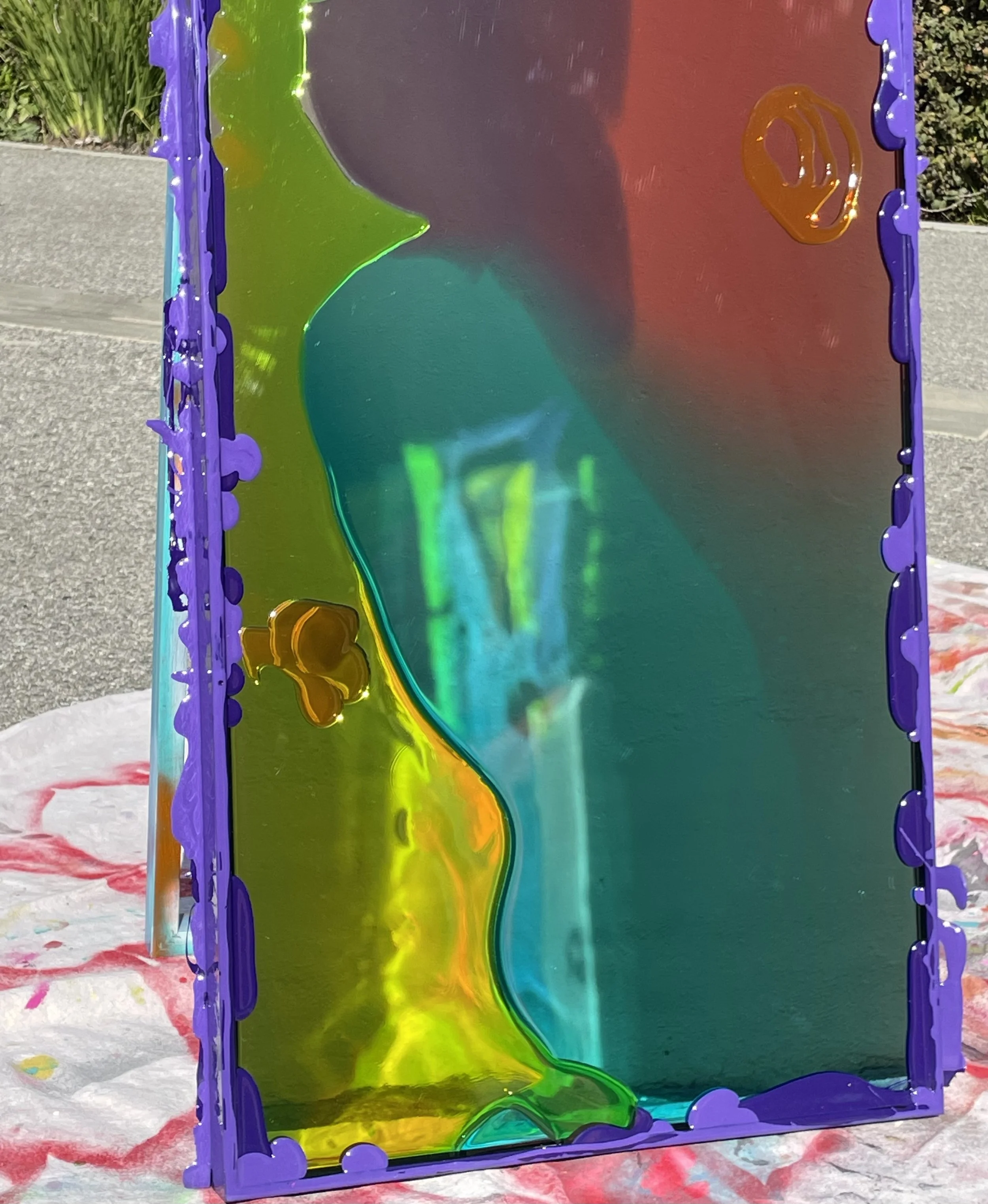 Colorful resin mirror art piece with purple border, reflective surface, and abstract shapes, outdoors on a sidewalk.