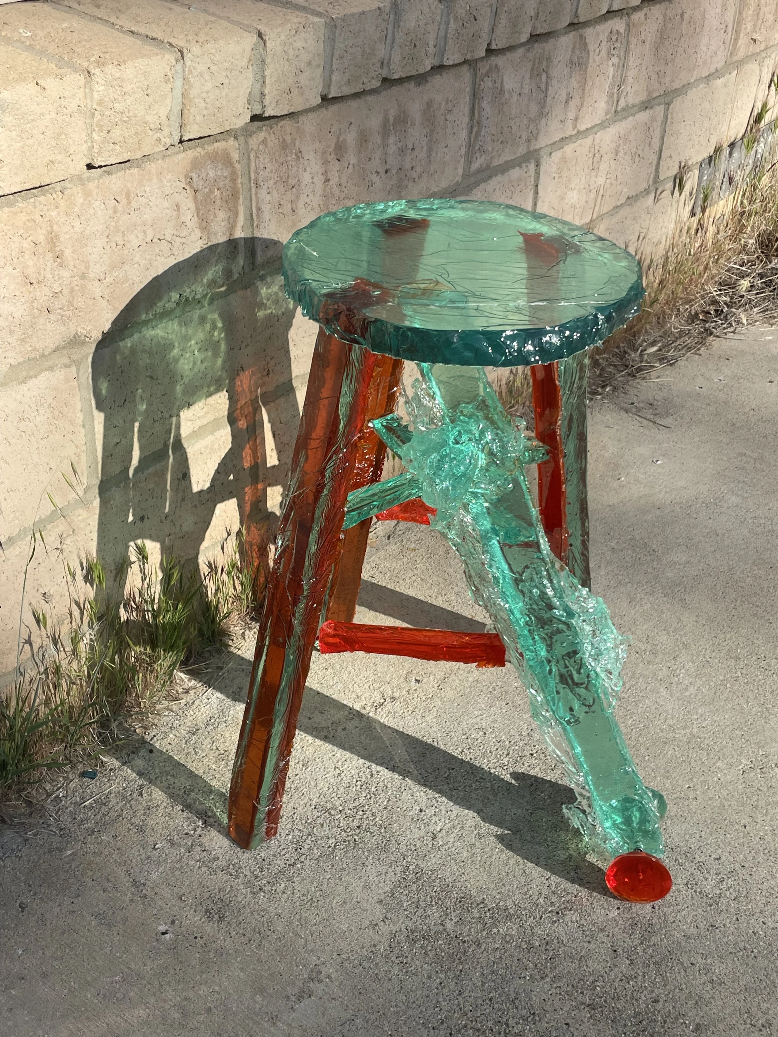 Glass art sculpture of a colorful high chair with a round seat, three legs, and a star-shaped backrest, placed outdoors against a brick wall.