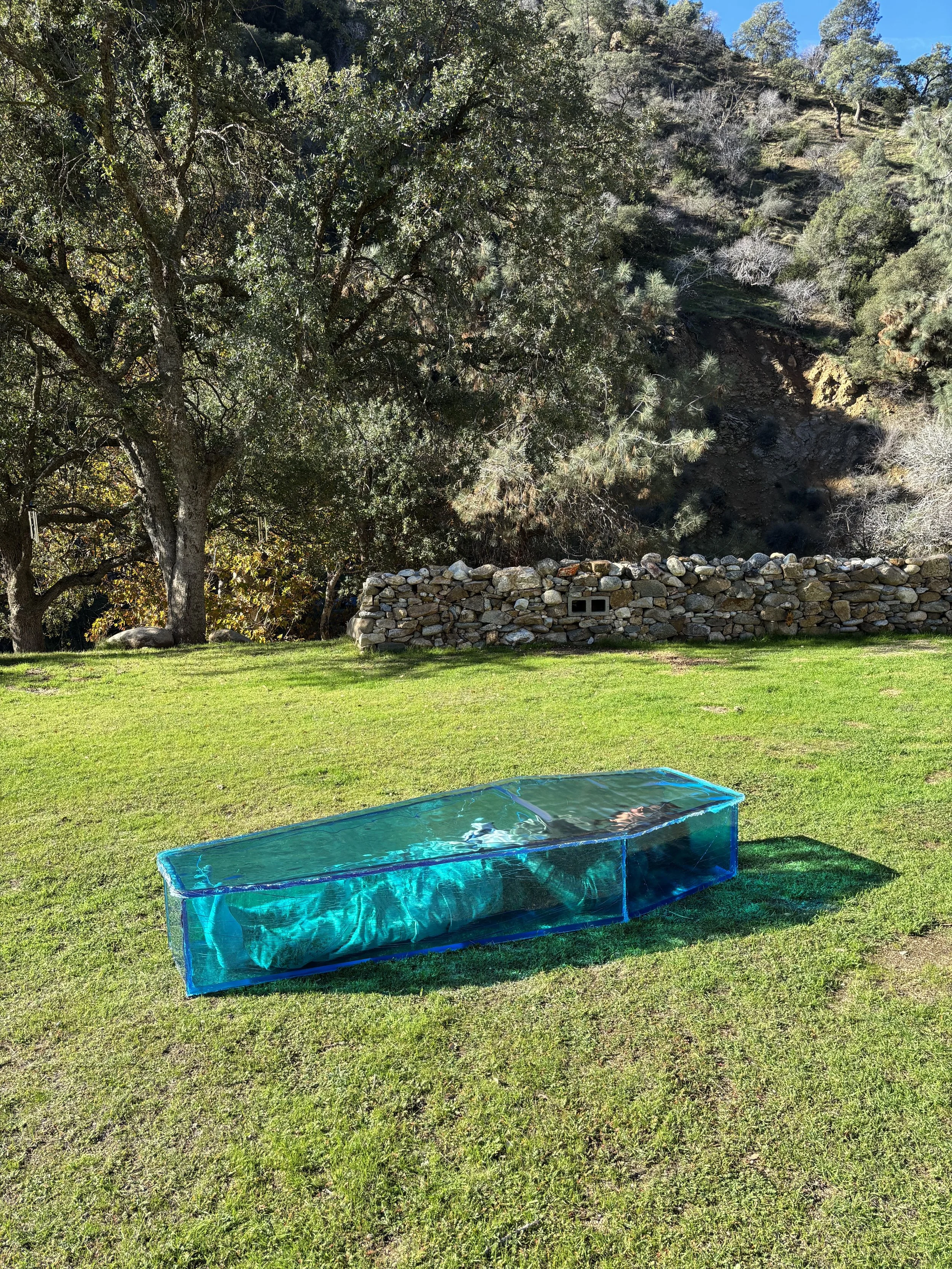 A transparent blue resin coffin placed on a grassy lawn, with trees and a stone wall in the background under a clear sky. Someone is inside of the coffin.