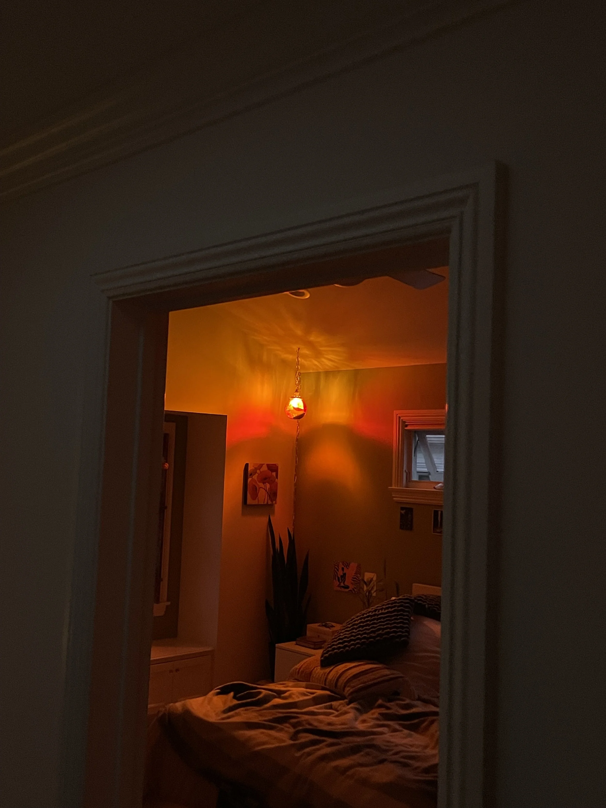 A bedroom illuminated by a colorful resin light, with a bed, pillows, a small window, a plant, and wall art.