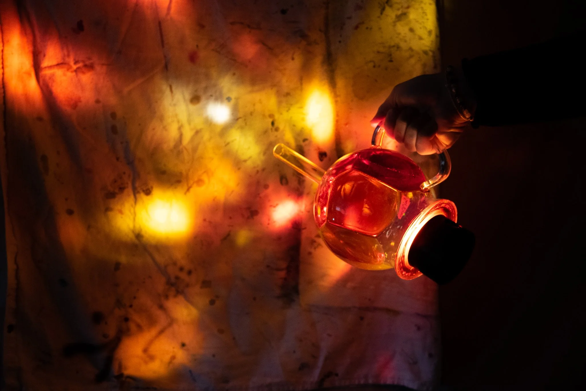 A hand holding a glass coffee siphon filled with orange and yellow resin over marbled backdrop with colorful light reflections.
