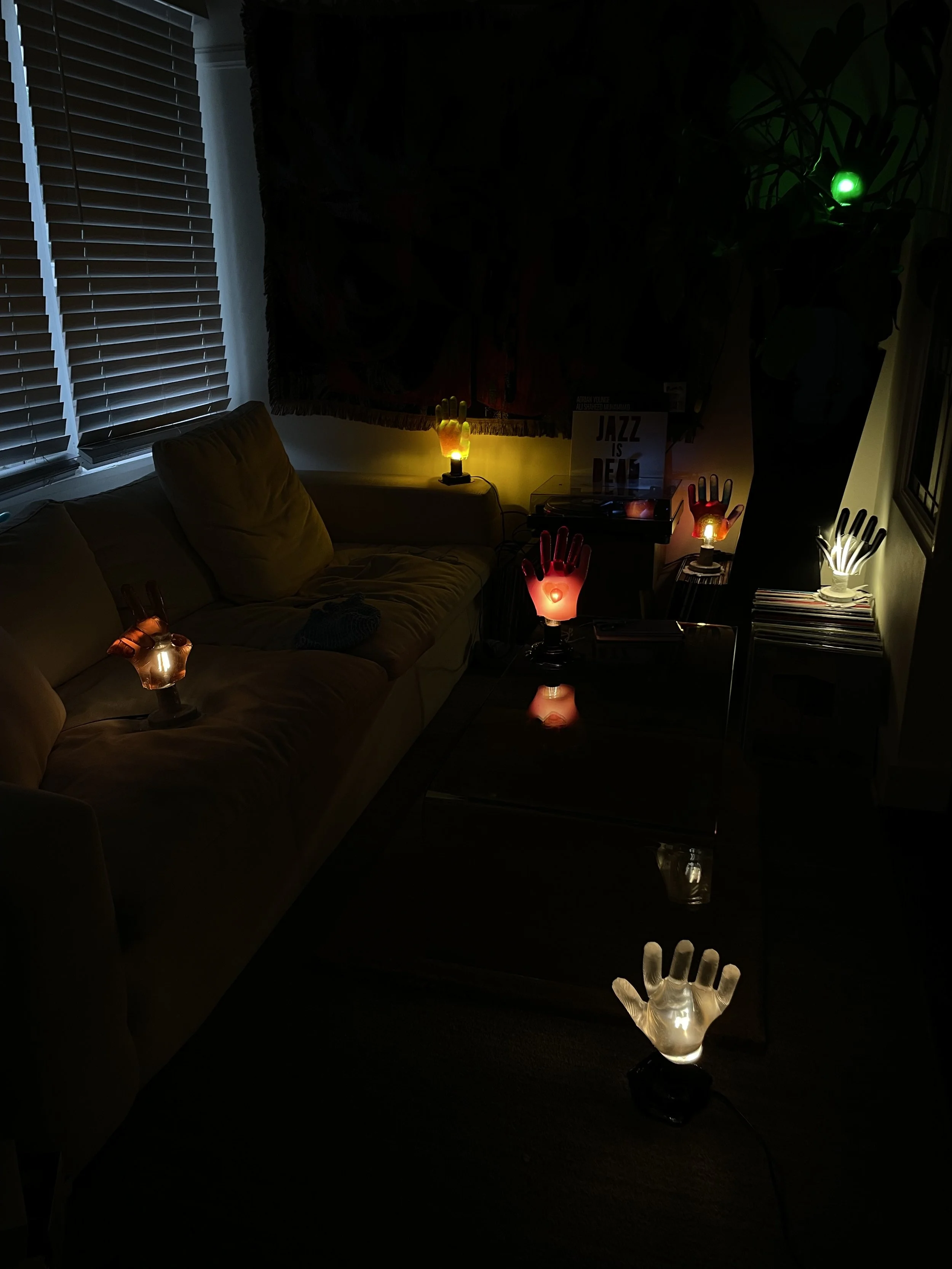 Living room with illuminated hand-shaped resin lamps of various colors on tables and furniture, partially lit by natural light from window blinds.