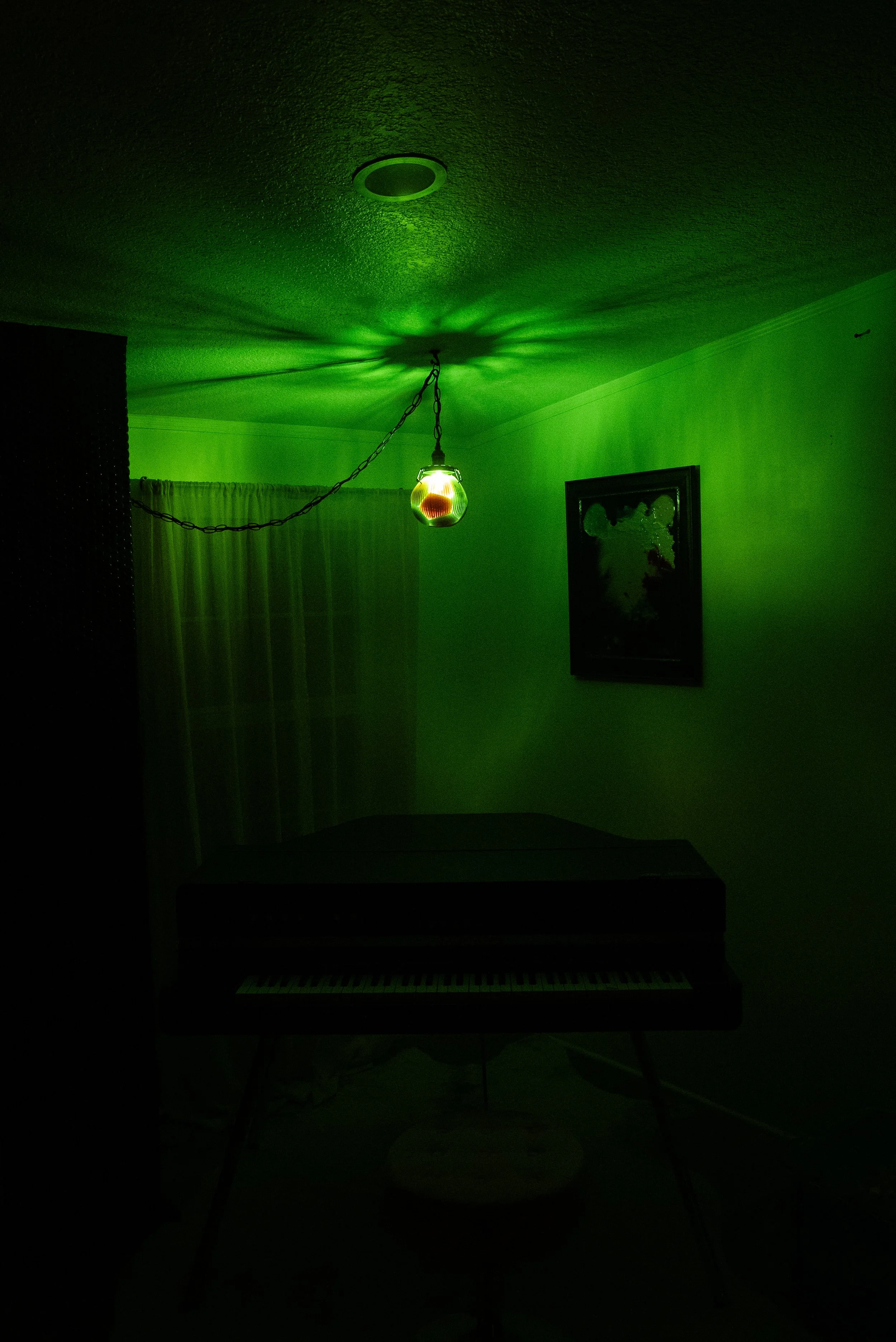 A dimly lit room with a green hue cast by a hanging, multi-colored resin light fixture. There is a piano against the wall, a framed picture, a curtain, and a dark cabinet.