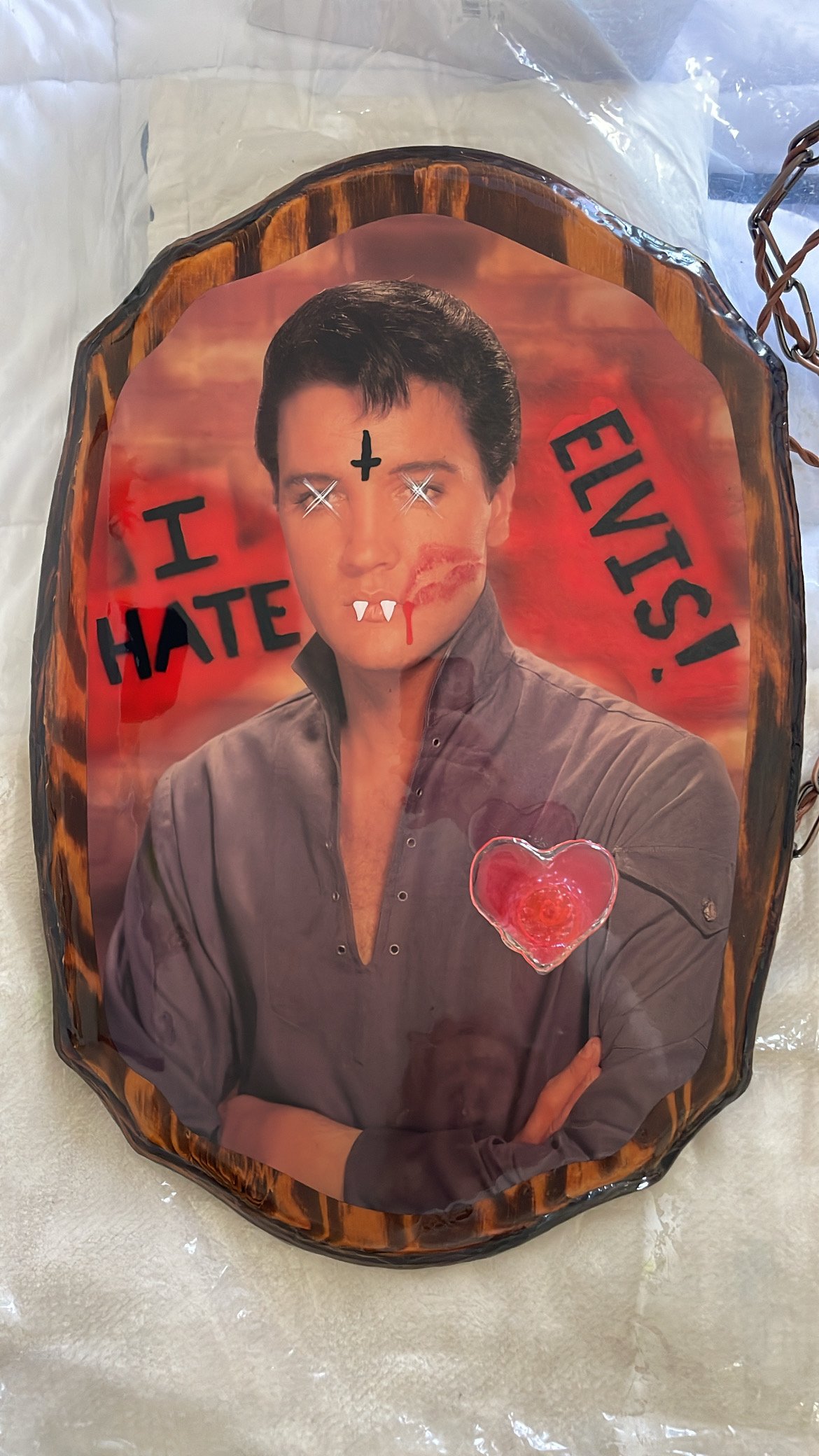 A decorative plaque featuring an edited image of Elvis with vampire fangs, a cross on his forehead, and scratched eyes. The words "I HATE ELVIS!" are written in bold black  letters. There's a heart-shaped resin light on his chest.