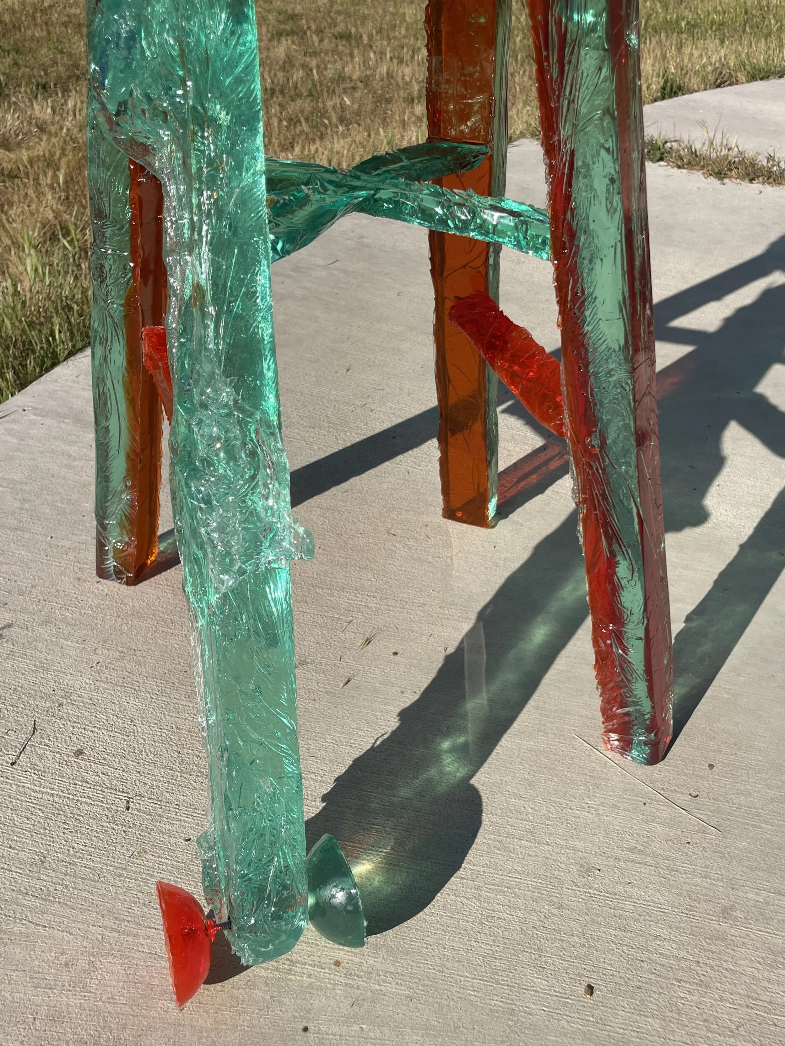 Close-up of a colorful glass-like resin sculpture with red, green, and orange glass pieces, casting shadows on a concrete surface outdoors.