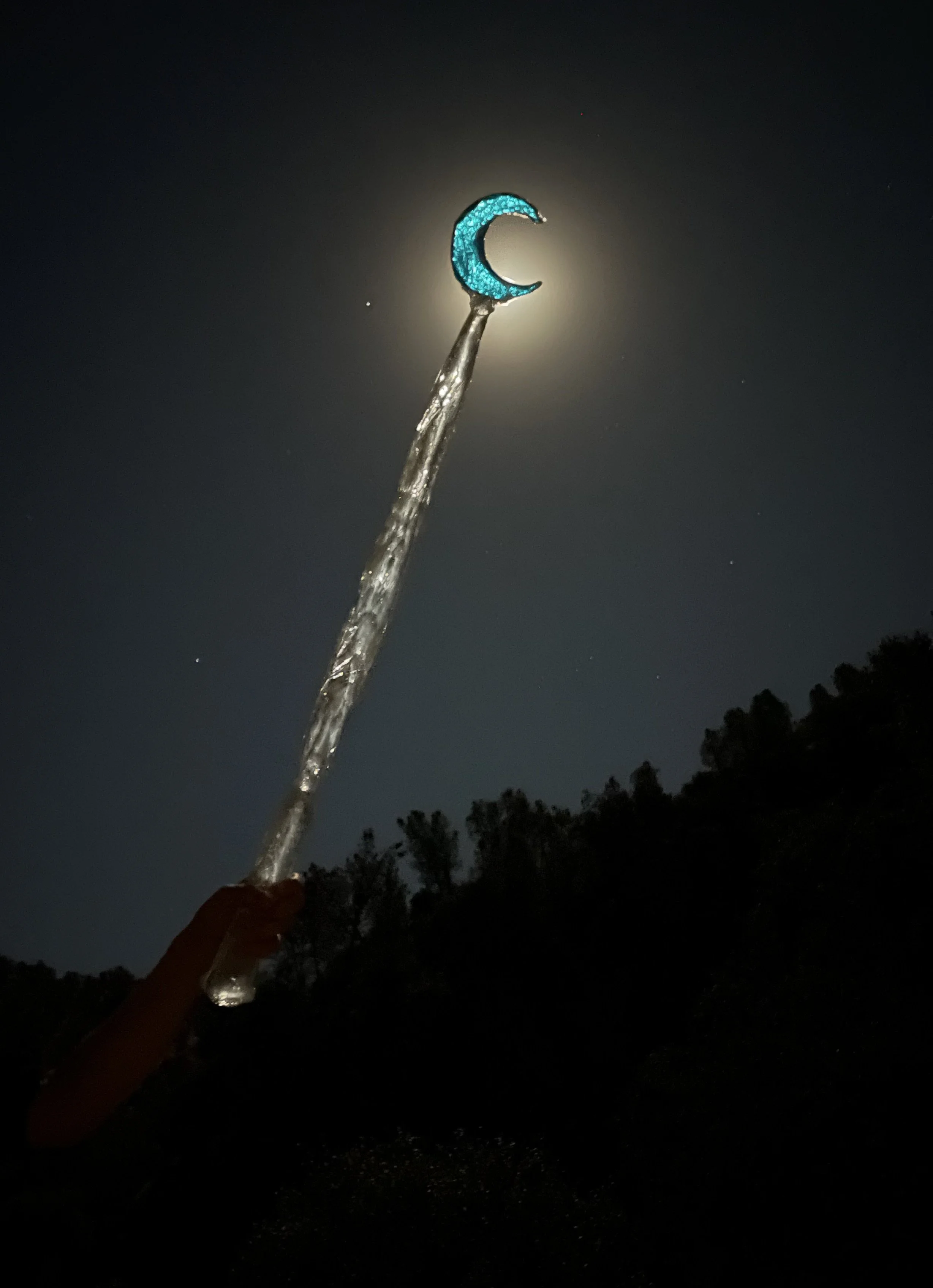 A person holding a resin magic wand with a glowing blue crescent moon at the tip against a night sky illuminated by the moon and stars.