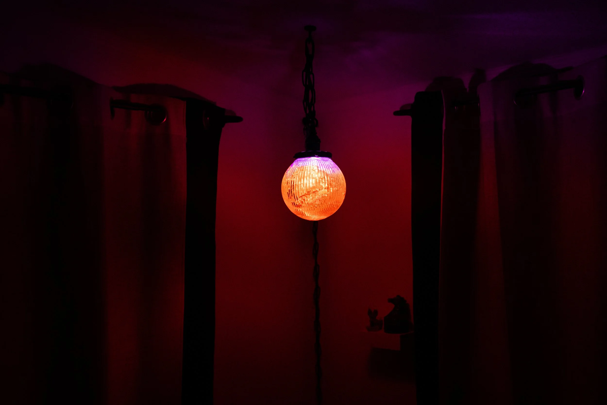A hanging resin lamp with an orange and purple light illuminating a dark room, with curtains and a small shelf with decor in the background.
